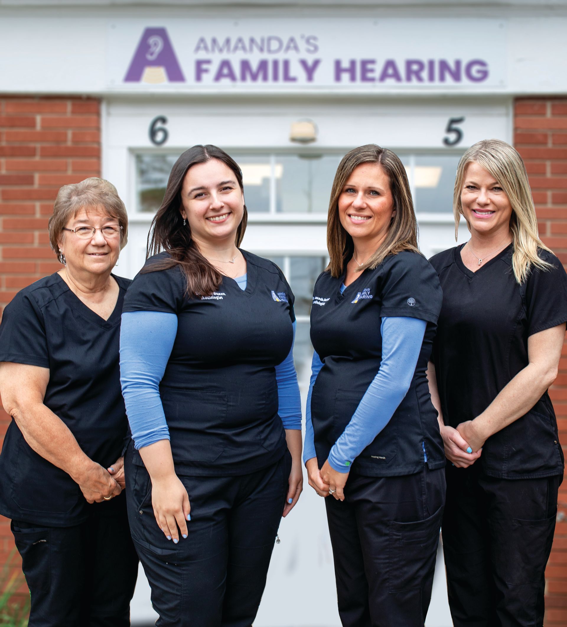 Four smiling employees in black scrubs standing in front of the Amanda’s Family Hearing office building.