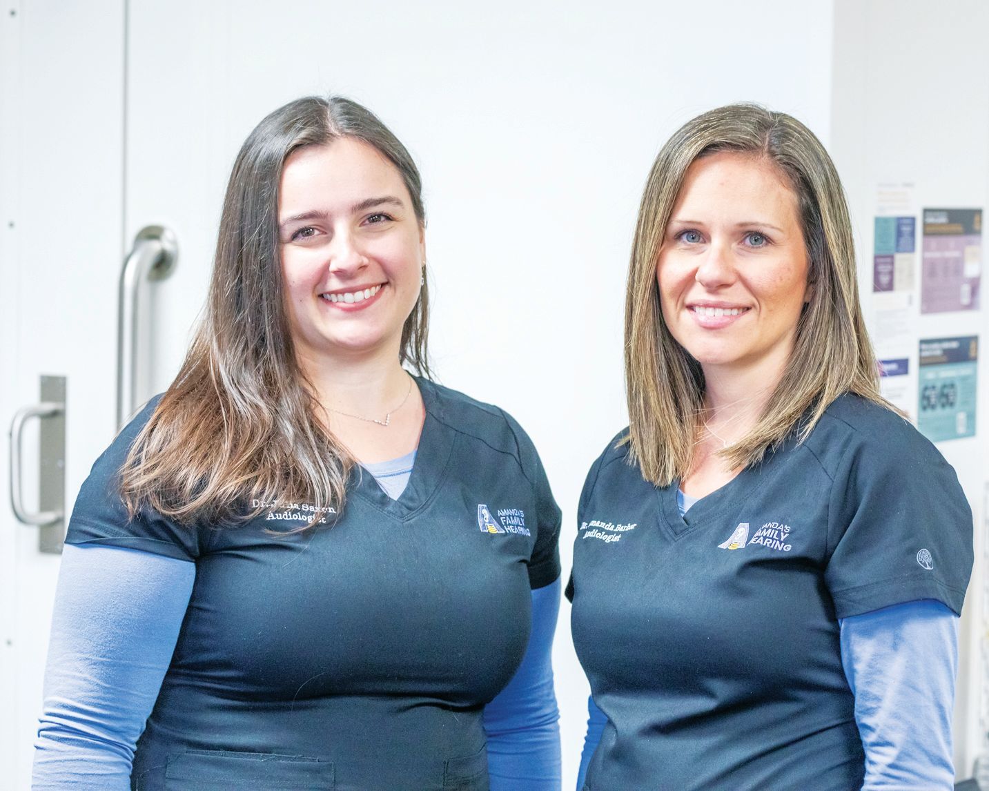 Two professionals in blue scrub tops and long-sleeved shirts standing side-by-side in a clinical office setting.