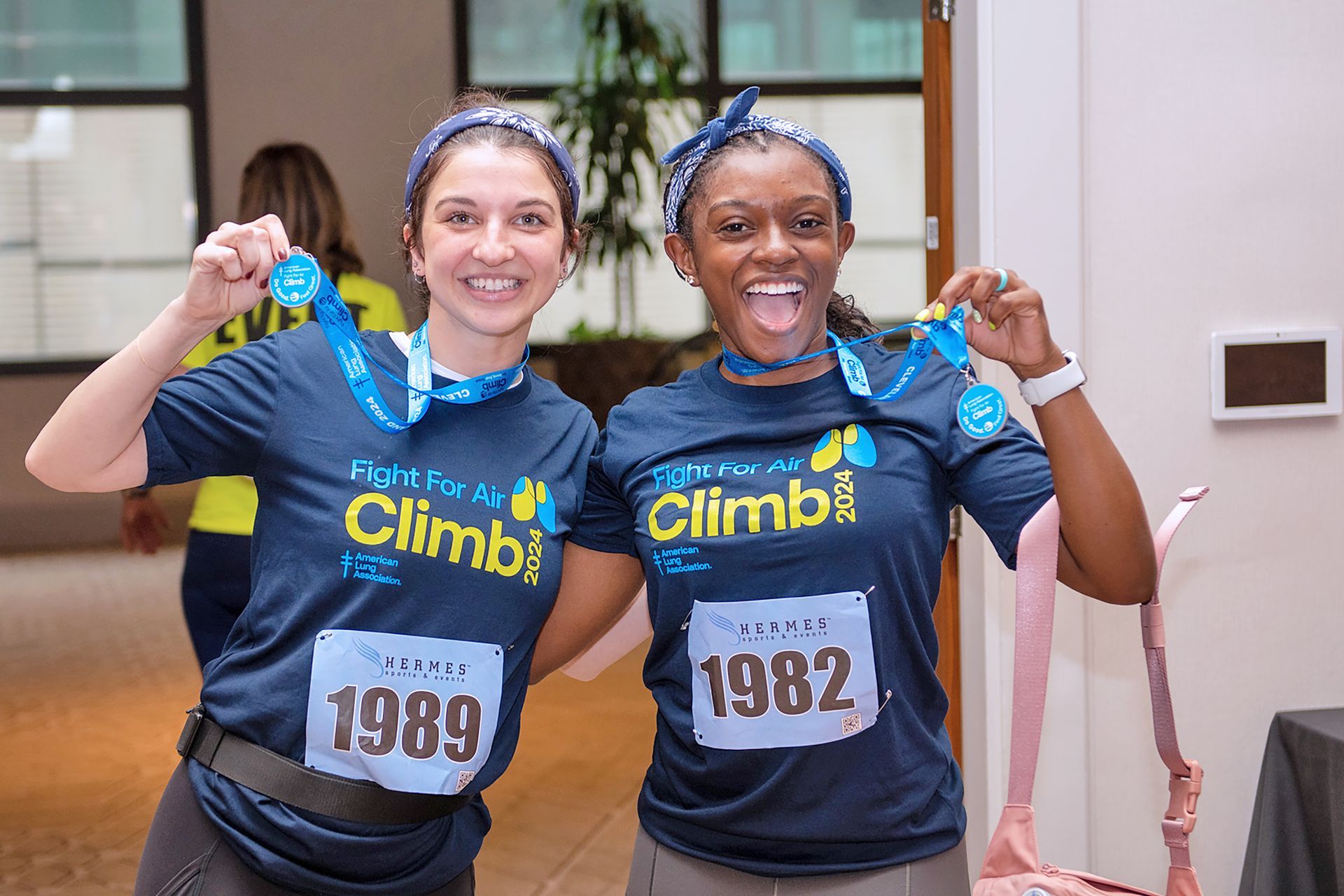 Two participants in matching navy event shirts and headbands hold up finisher medals, smiling at an indoor event.