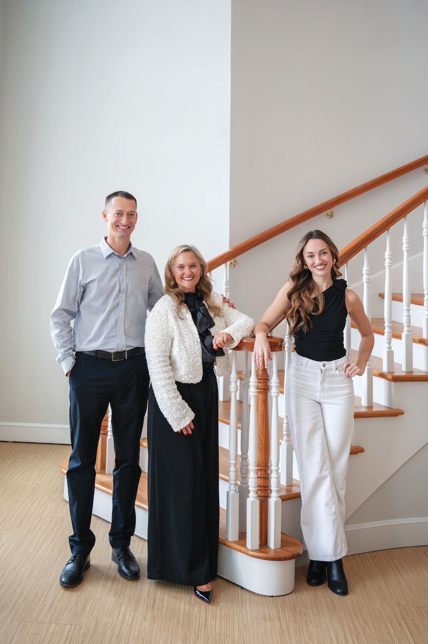 Three people pose smiling on the stairs of a home, wearing casual to dressy attire.