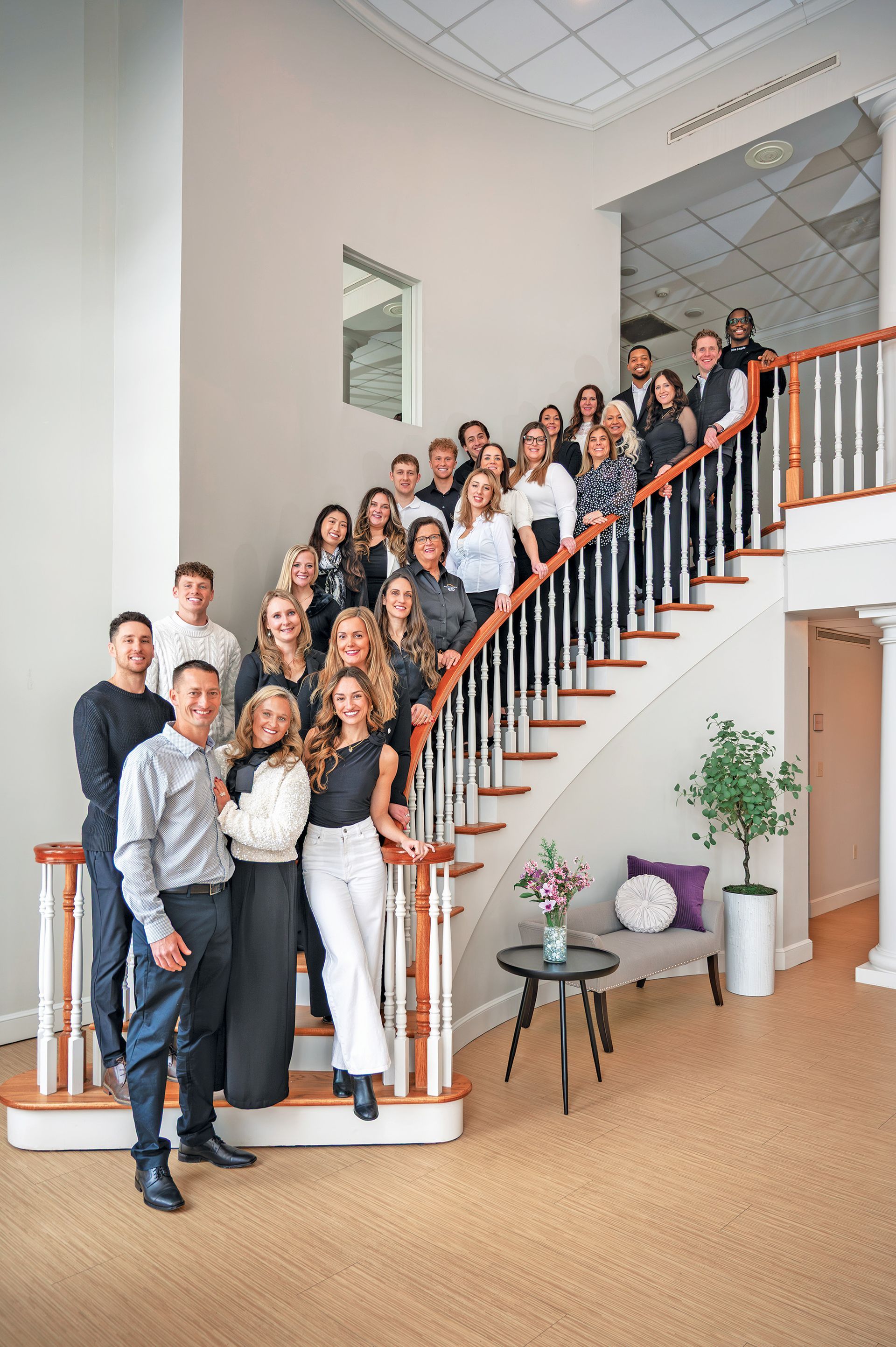 A large group stands on a staircase in a bright, modern foyer, posed for a formal group portrait.