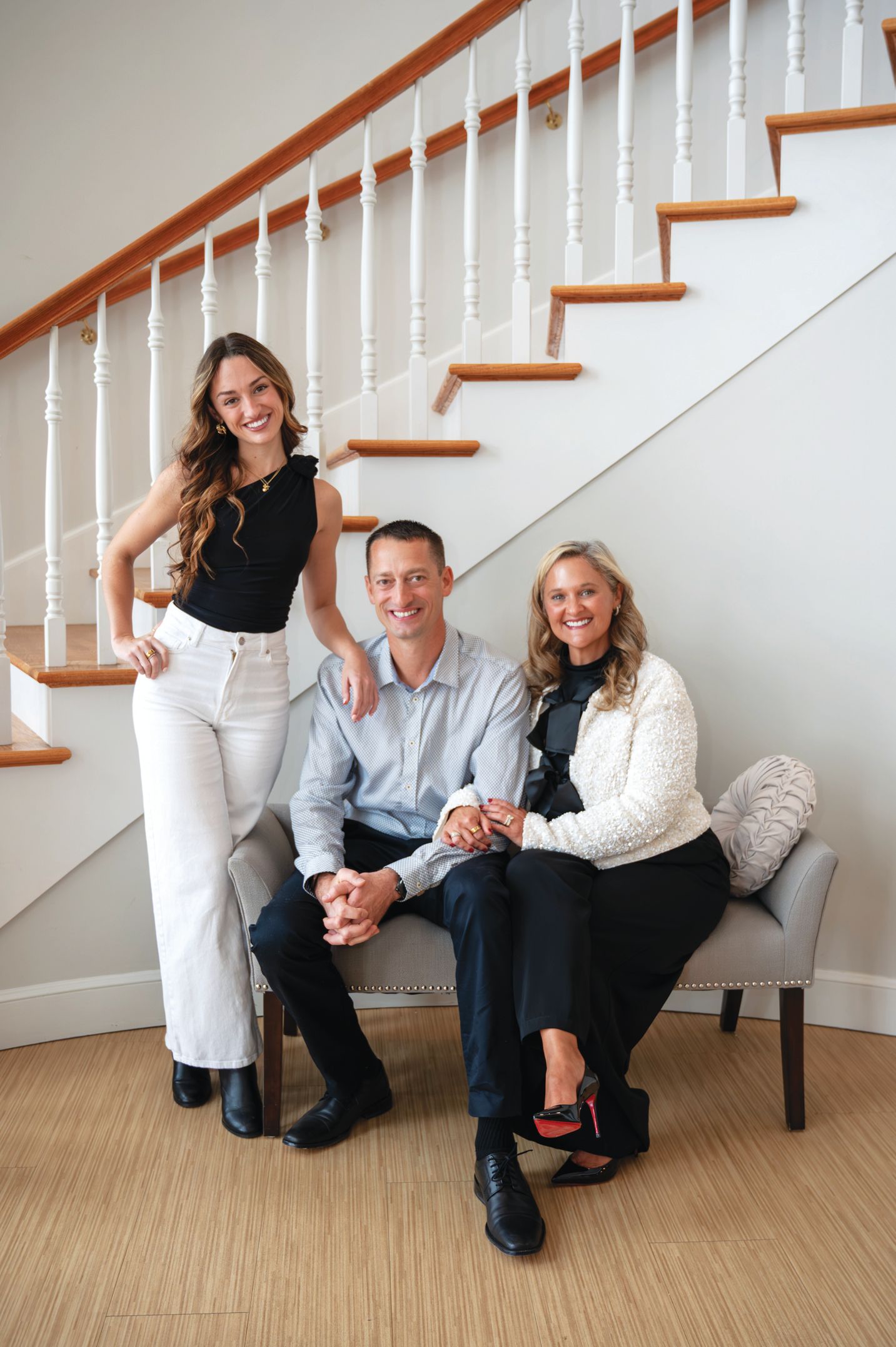 A family of three poses in front of a white staircase; one person stands while two sit on a bench, all smiling warmly.