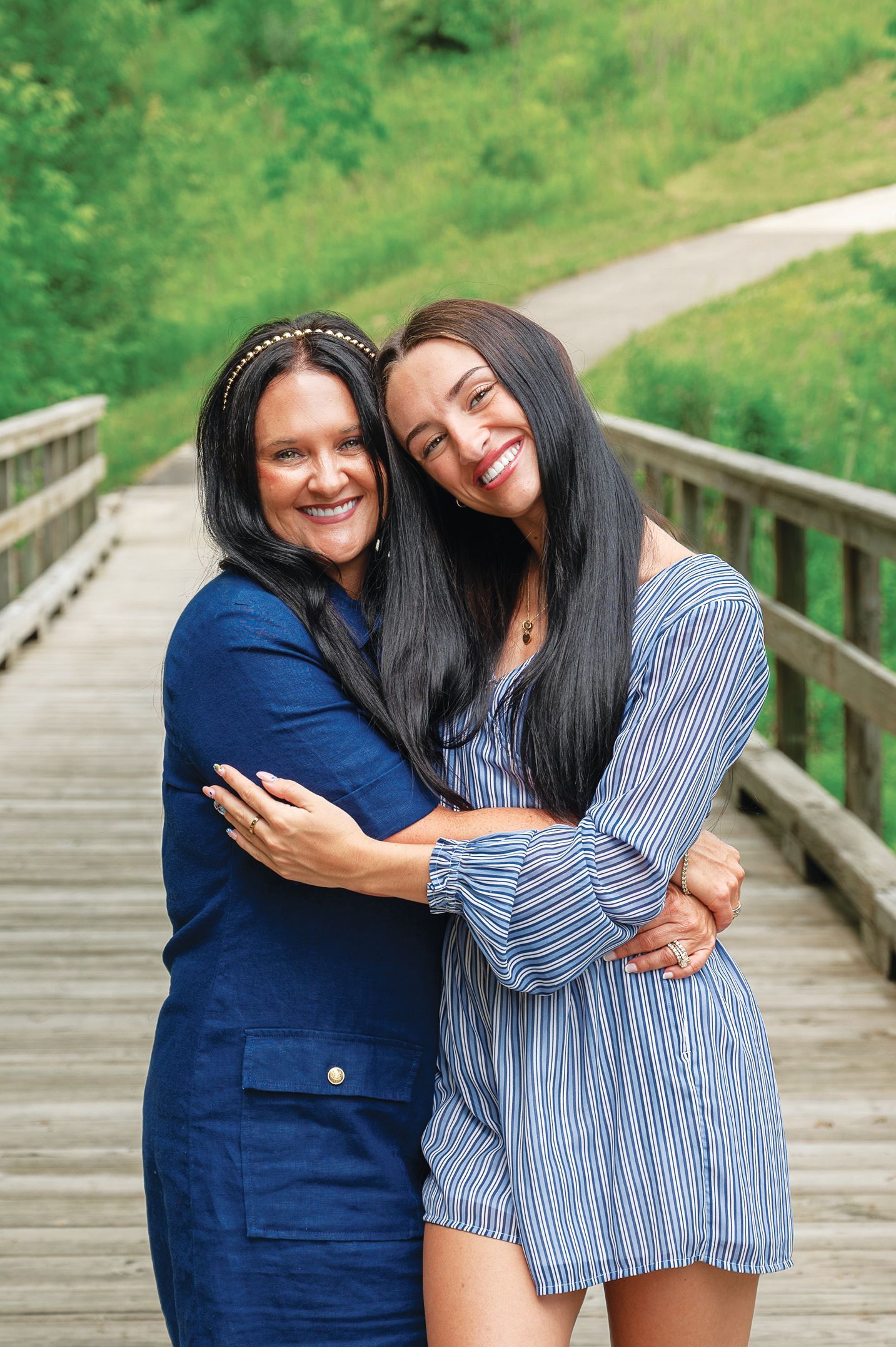 Two smiling individuals embrace on a wooden bridge set against a grassy, outdoor background.