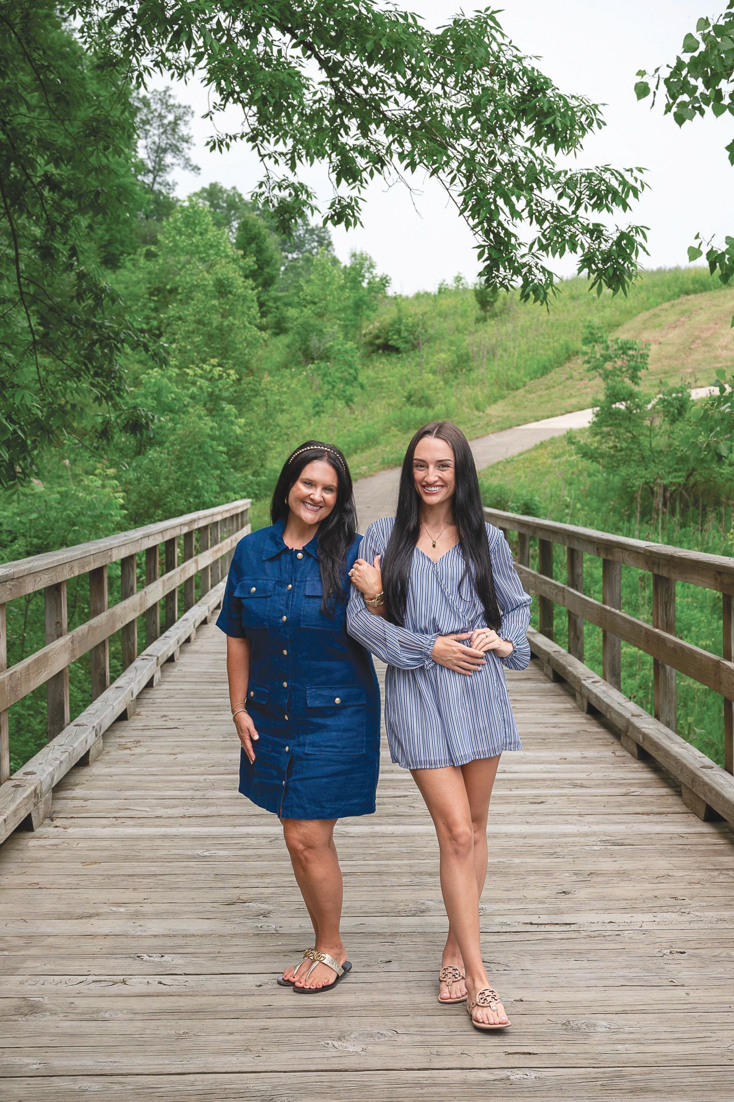 Two people standing arm-in-arm on a wooden bridge outdoors, surrounded by lush green trees and a grass-covered hill.