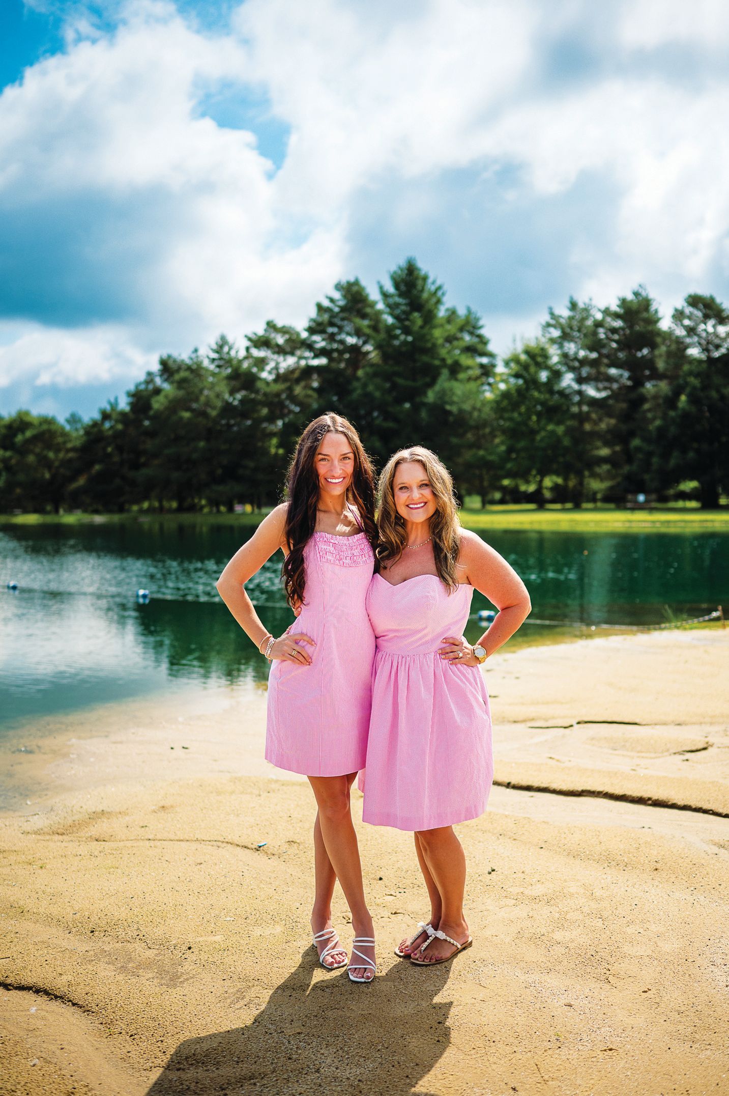Two individuals in matching pink dresses smile while standing on a sandy beach by a lake in front of a forest.