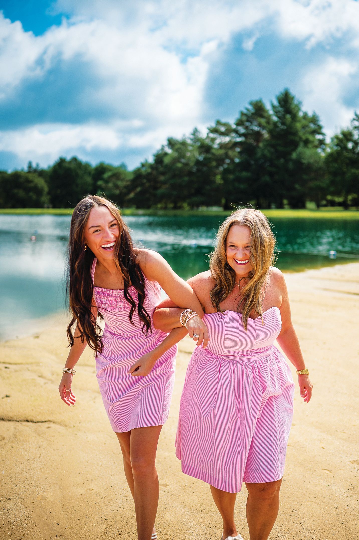 Two people in matching pink and white patterned dresses laughing while walking along a sandy beach by a lake.