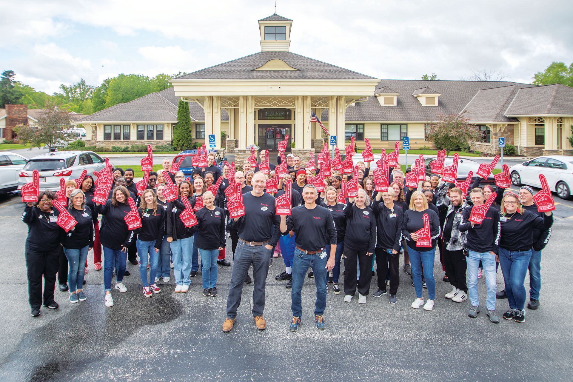 A large group of people in black shirts stands outside a building, all holding red foam fingers toward the camera.