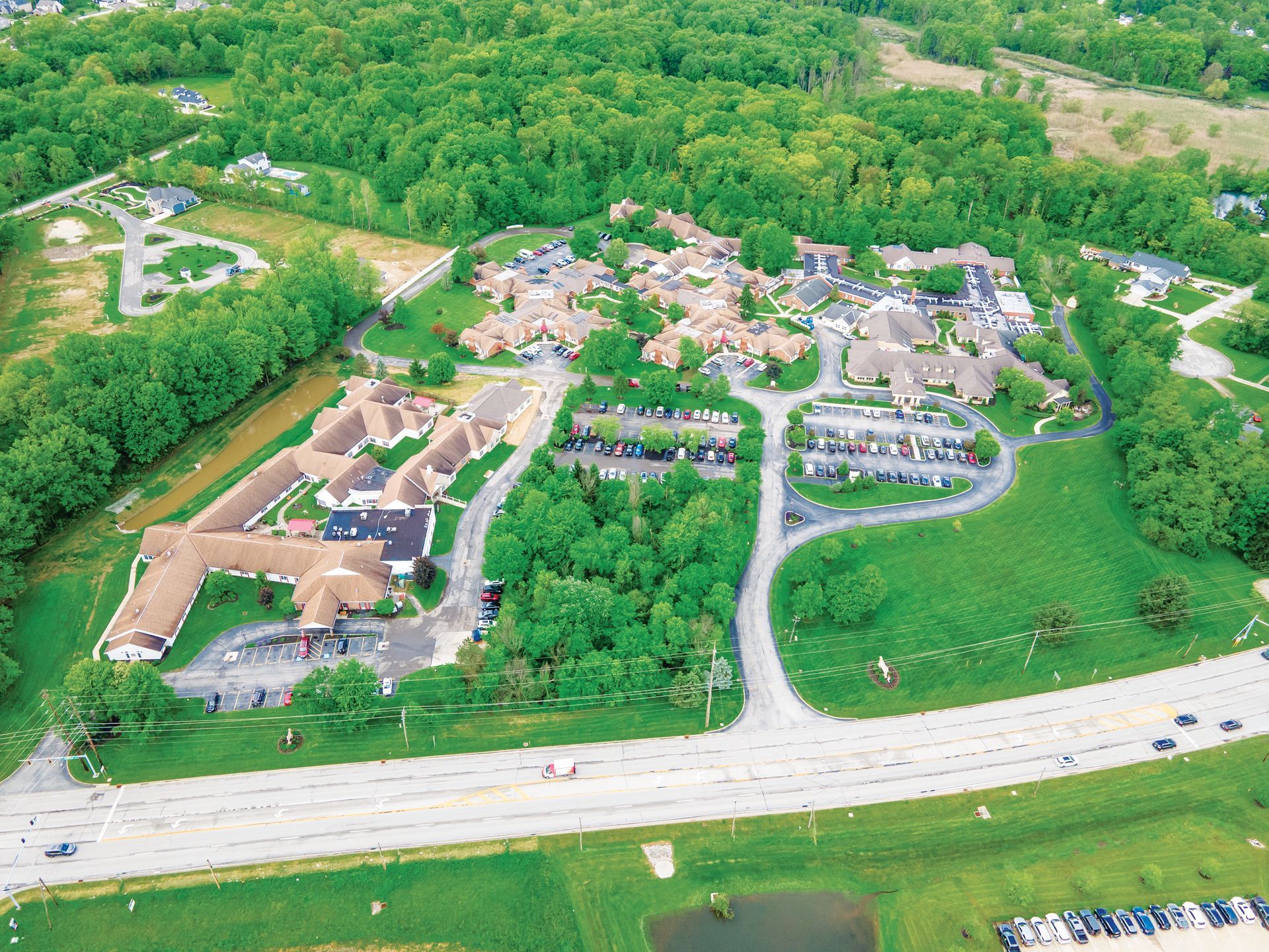 An aerial view of a multi-building residential or care facility complex surrounded by trees, grass, and parking lots.