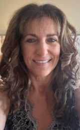 A smiling woman with long, curly, brown hair wearing a dark, lace-trimmed top.