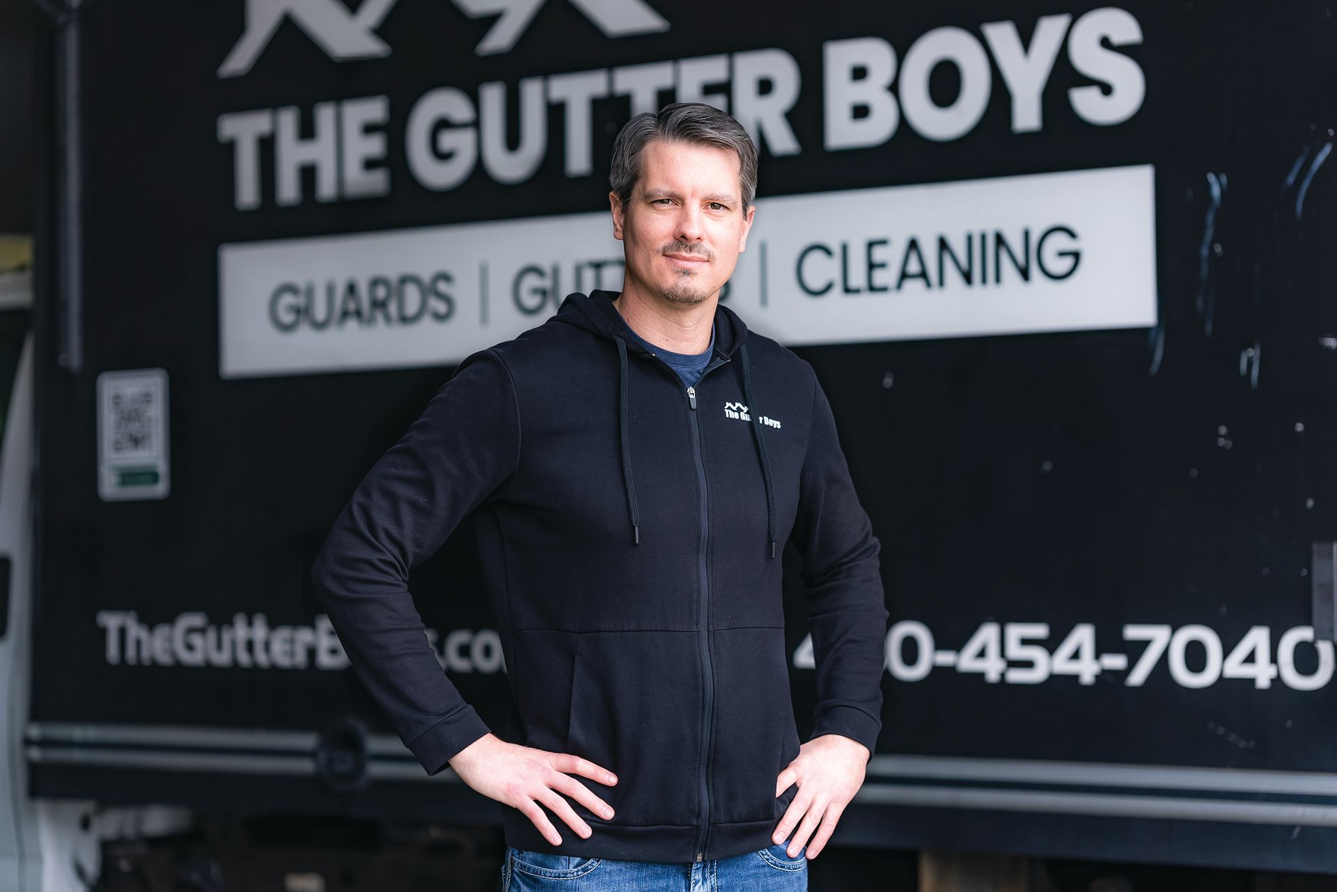 A person in a black hoodie stands with hands on hips in front of a service truck labeled 