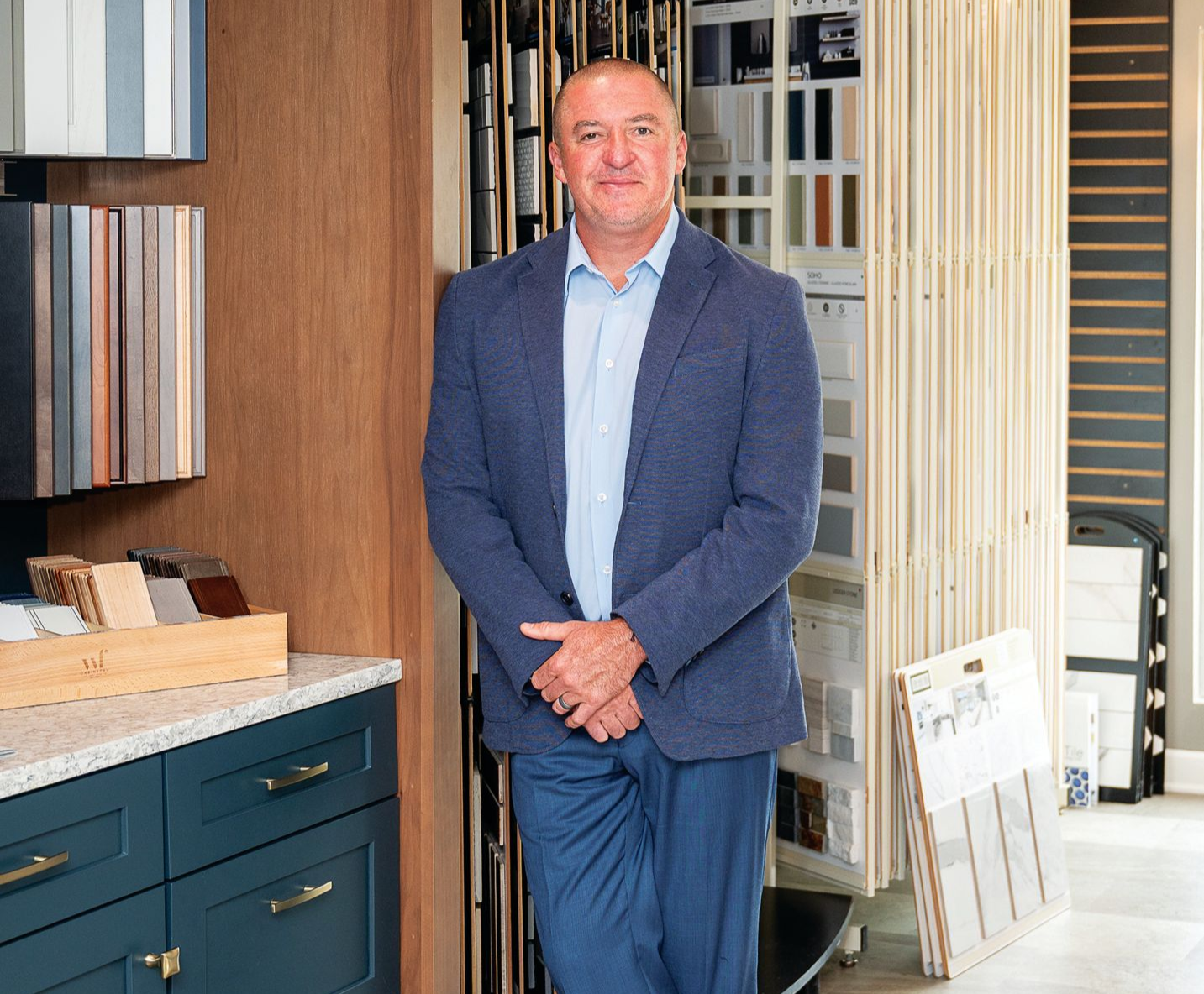A professional in a blue blazer and light shirt poses in a showroom with wall-mounted material samples and blue cabinets.