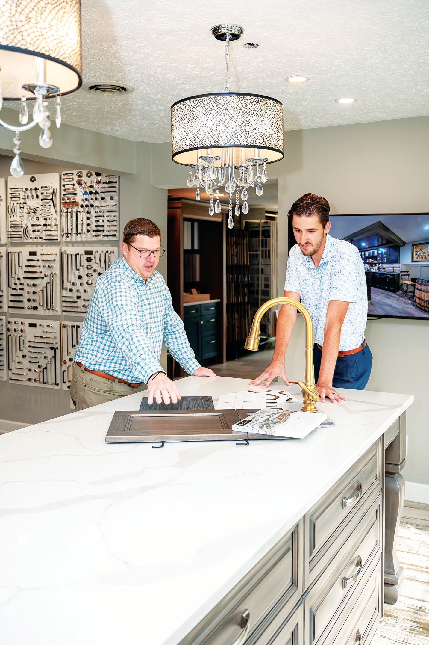 Two people reviewing material samples and a gold faucet on a large white marble kitchen island in a showroom.