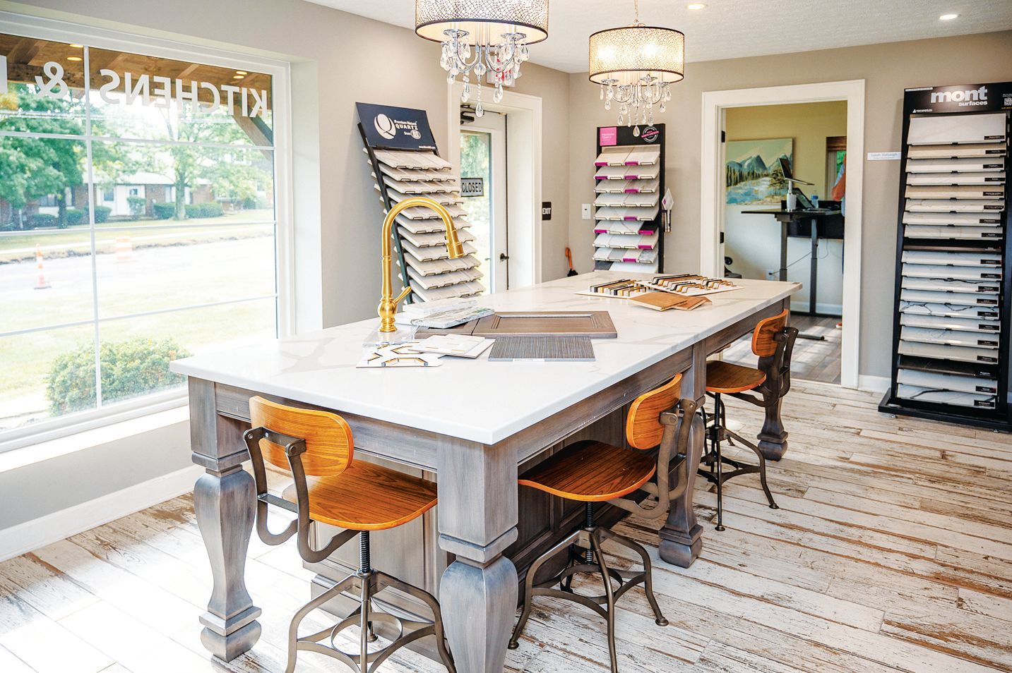 A bright showroom featuring a large center table with a stone top, surrounded by stools, display racks, and chandeliers.