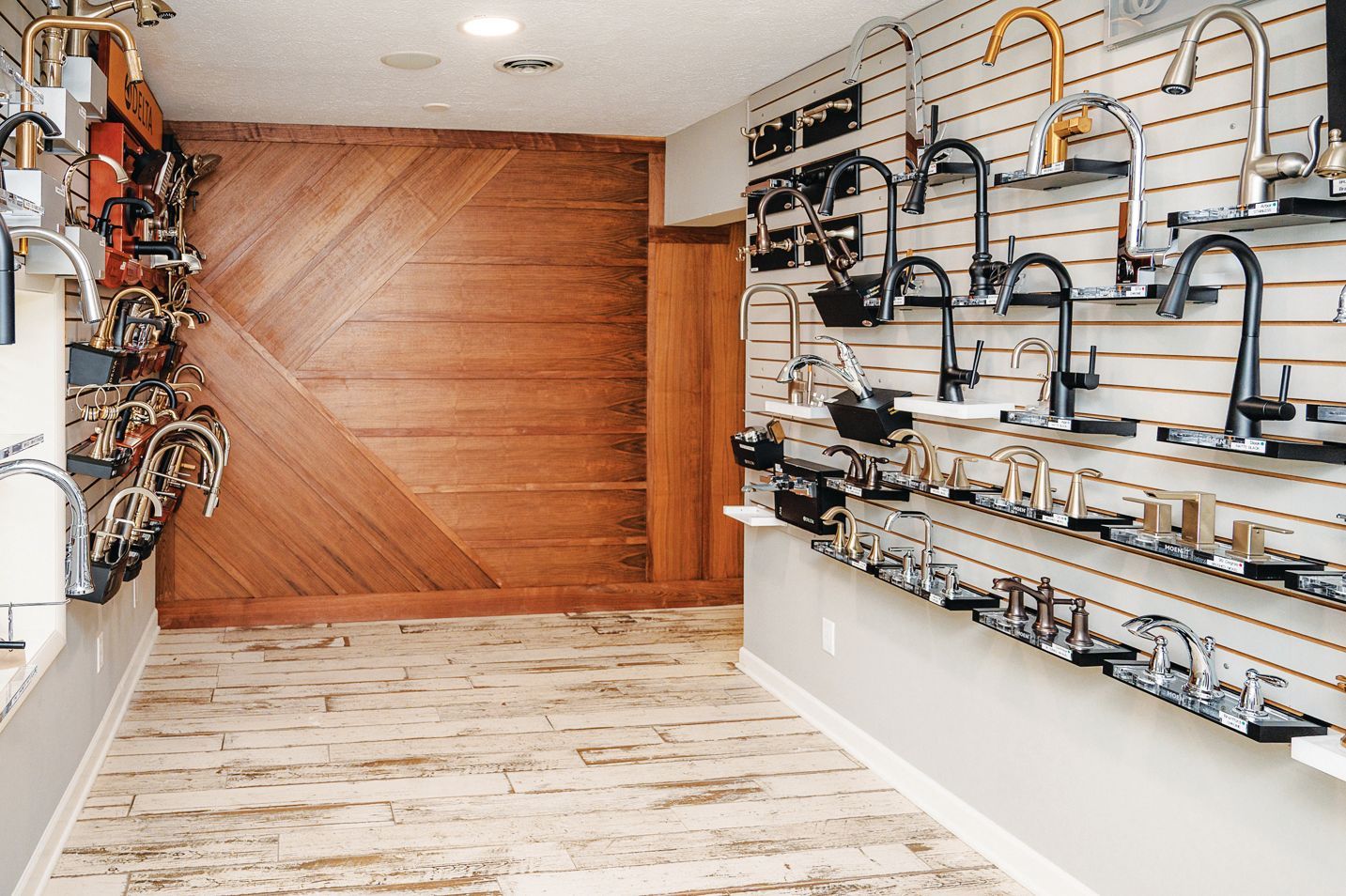 A showroom wall displays various kitchen and bathroom faucets on shelving against a wood-paneled feature wall.
