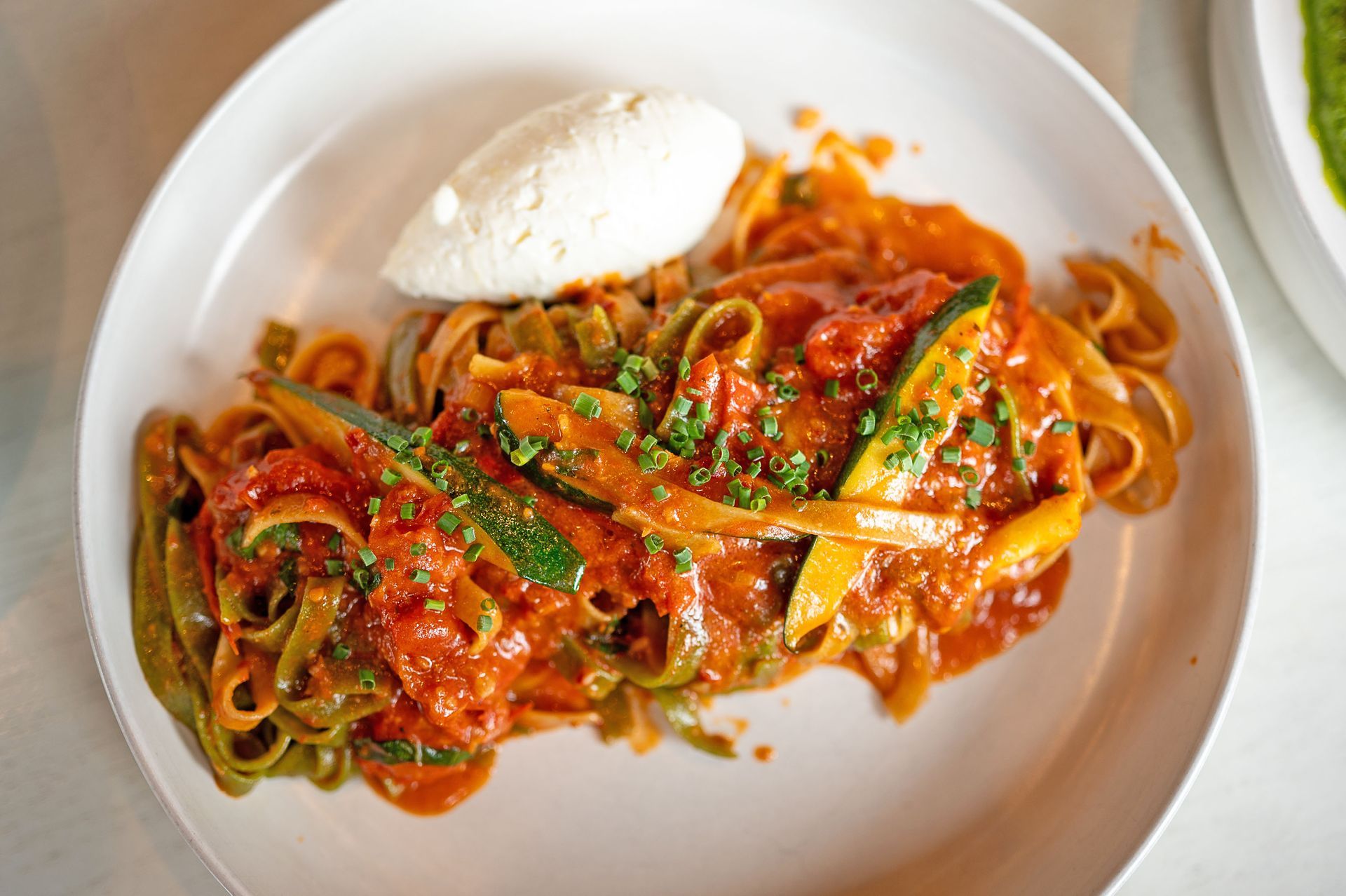 A plate of pasta tossed in tomato sauce with sliced zucchini and topped with a scoop of creamy white ricotta cheese.