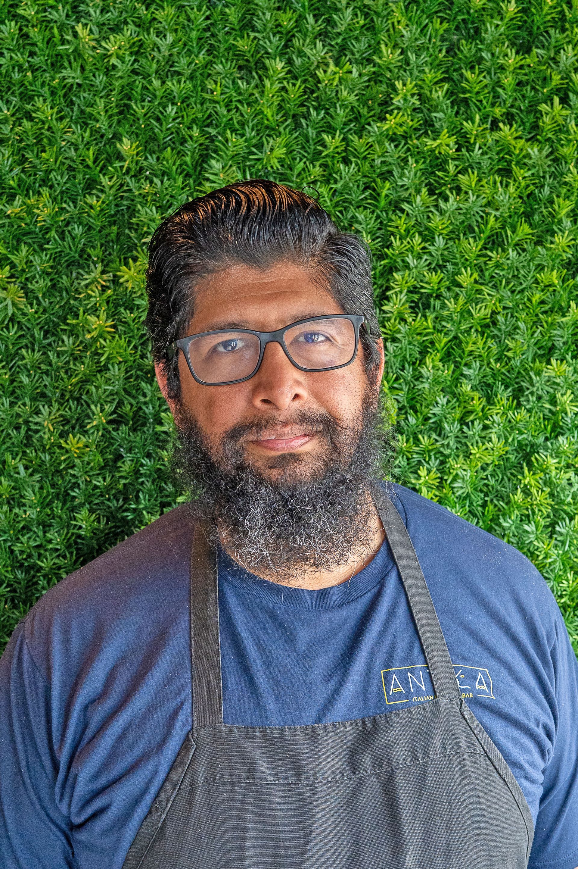 A person with a full beard and glasses, wearing a blue shirt and gray apron, poses in front of a green leafy backdrop.