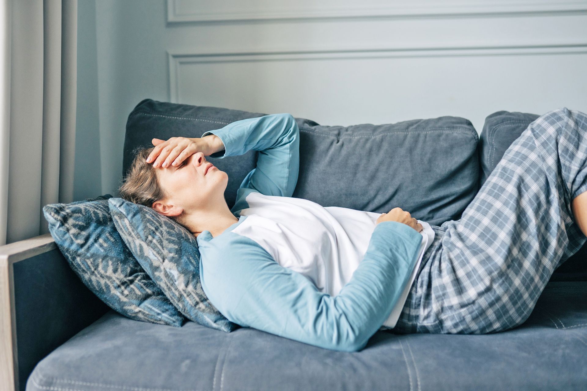 A person in light blue pajamas rests on a gray sofa with their hand over their eyes, appearing tired or in discomfort.