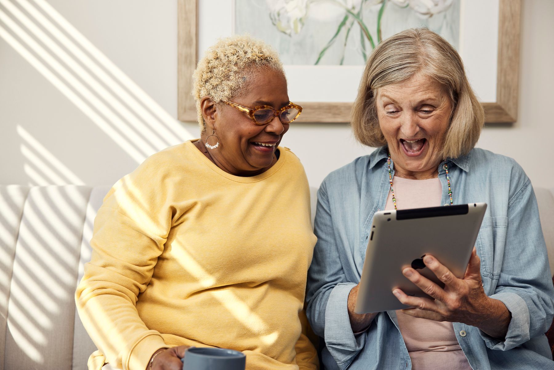 Two people sit side-by-side, smiling with excitement as they look at a tablet together in a sunlit room.