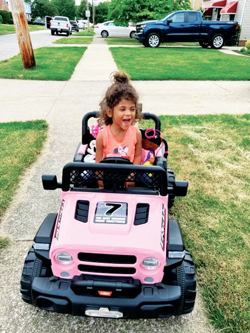 A young person with a joyful expression sits in a pink toy Jeep on a suburban sidewalk.