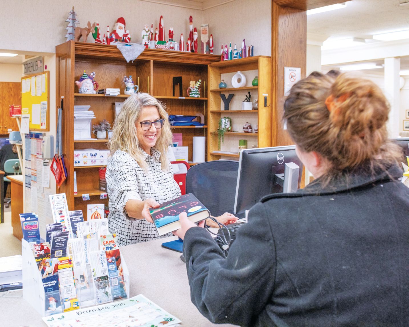 A person hands a book to another person at a library or office desk, with shelves filled with holiday decor in the back.