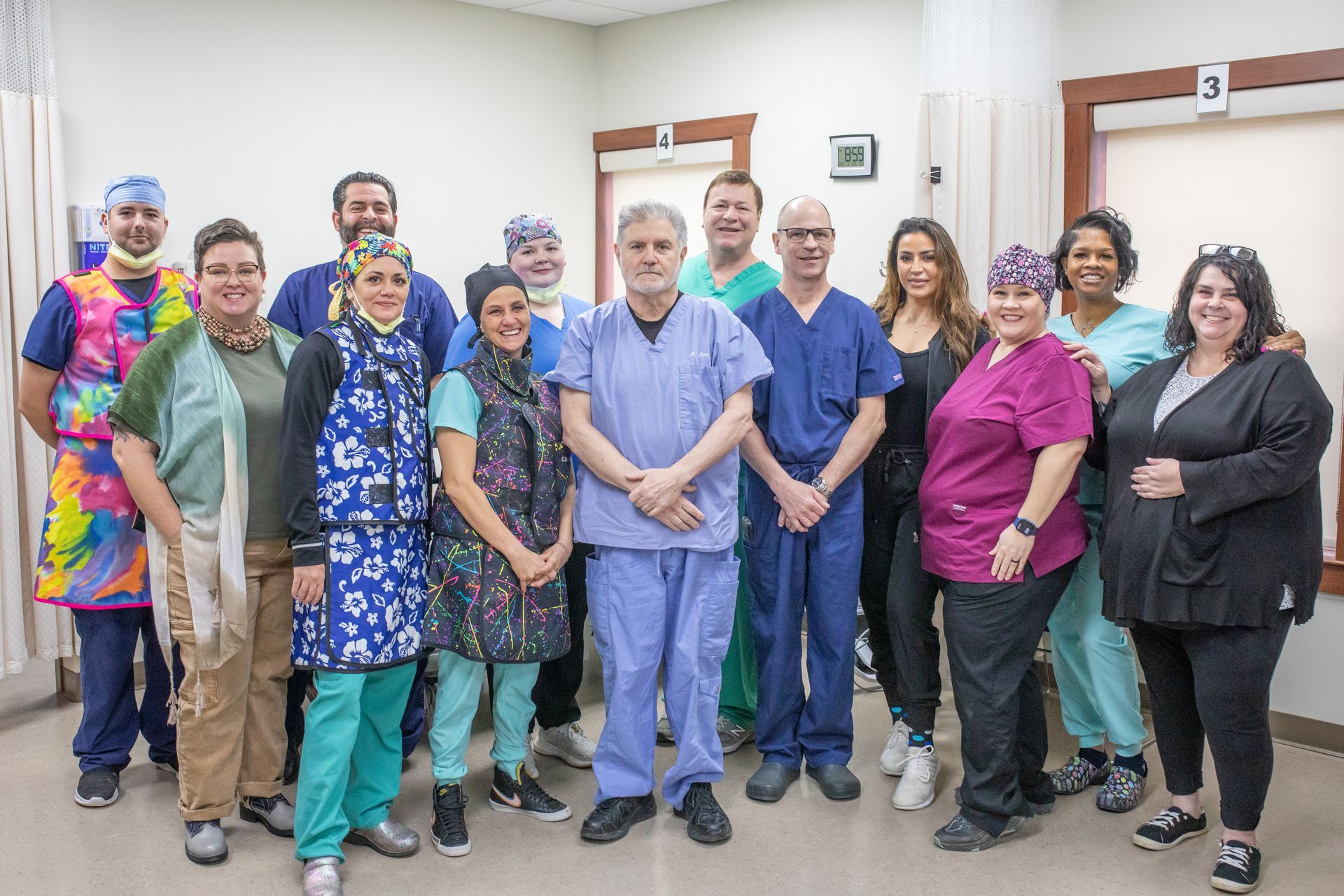A group of medical professionals in scrubs, vests, and casual attire stand together smiling in a clinical setting.