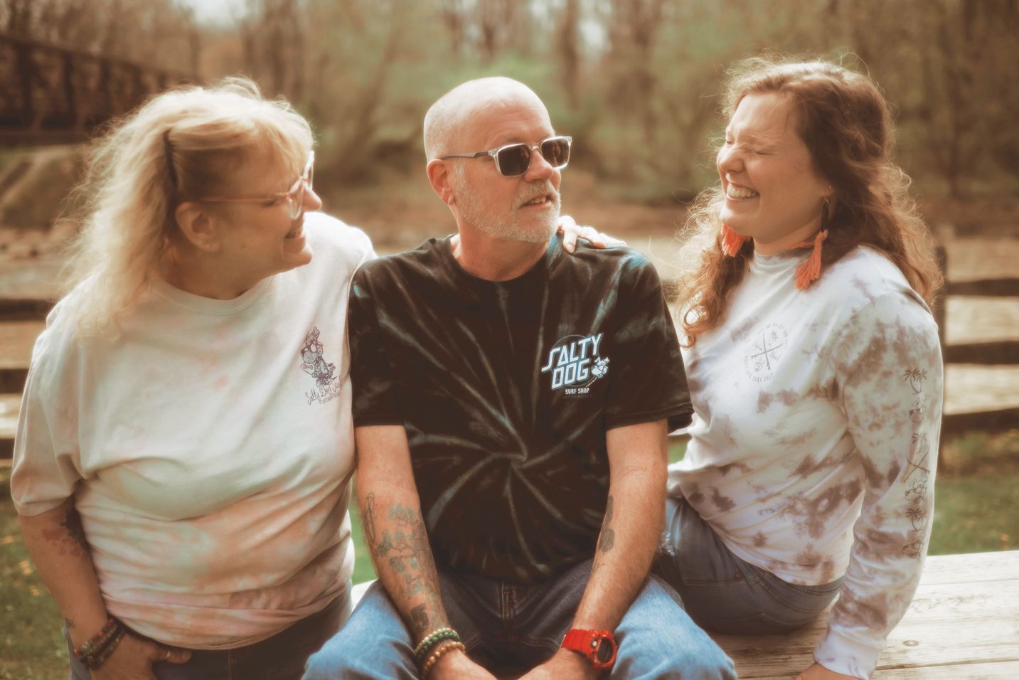 Three people sit on a wooden surface outdoors, smiling and looking toward each other in a casual, warm setting.