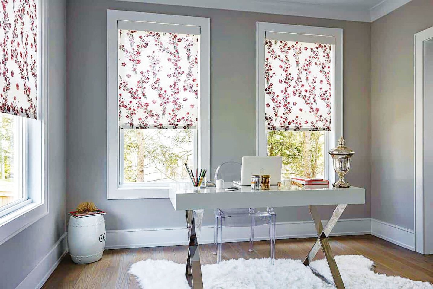 A bright home office with a desk, clear chair, and two windows covered in patterned floral roller shades.