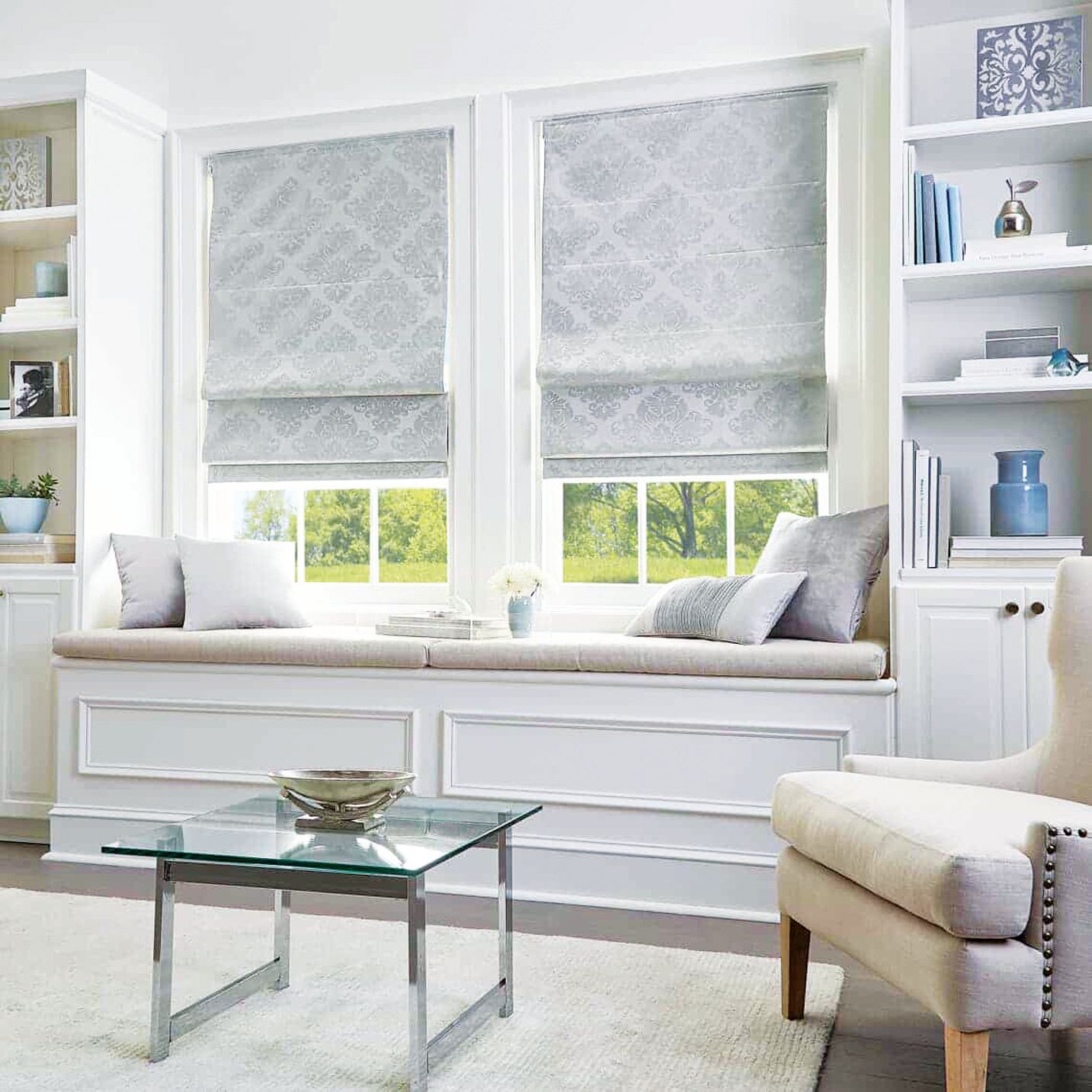 A white window seat with gray roman shades, throw pillows, a glass coffee table, and built-in white bookshelves.
