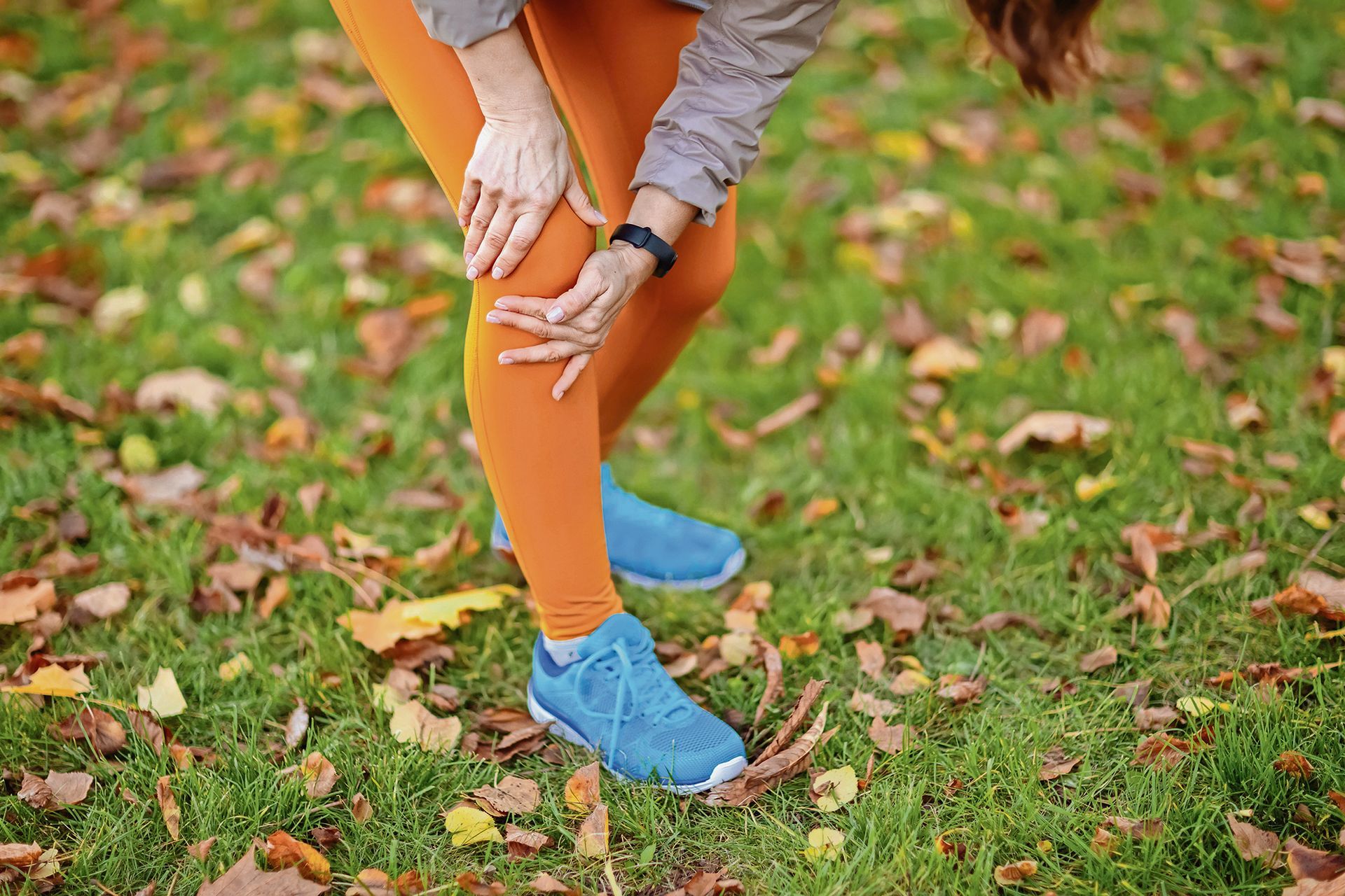 Person in orange leggings clutching their knee while standing in a park covered in autumn leaves.