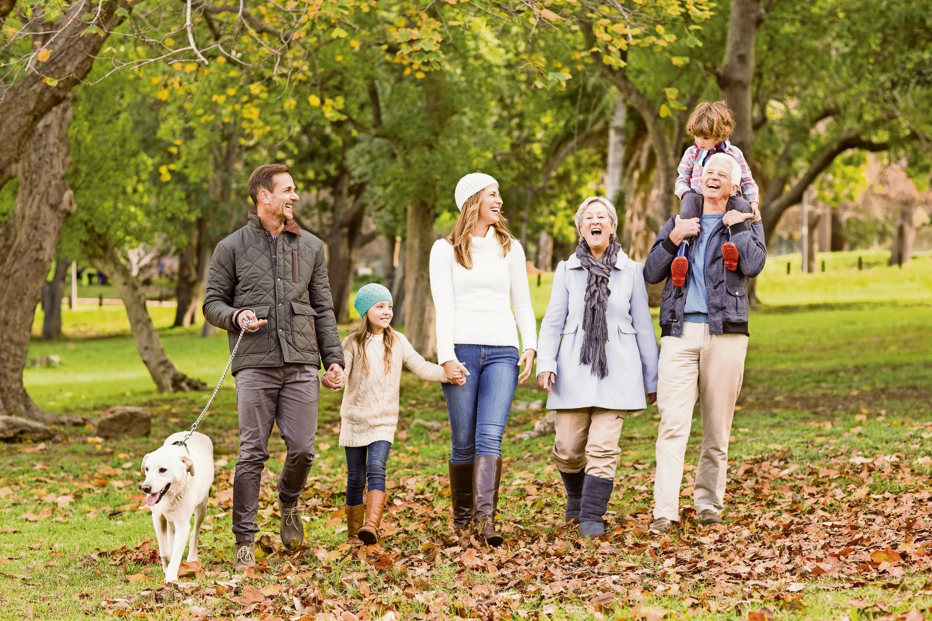 A family walks together through a park covered in autumn leaves, laughing while one adult carries a child on shoulders.