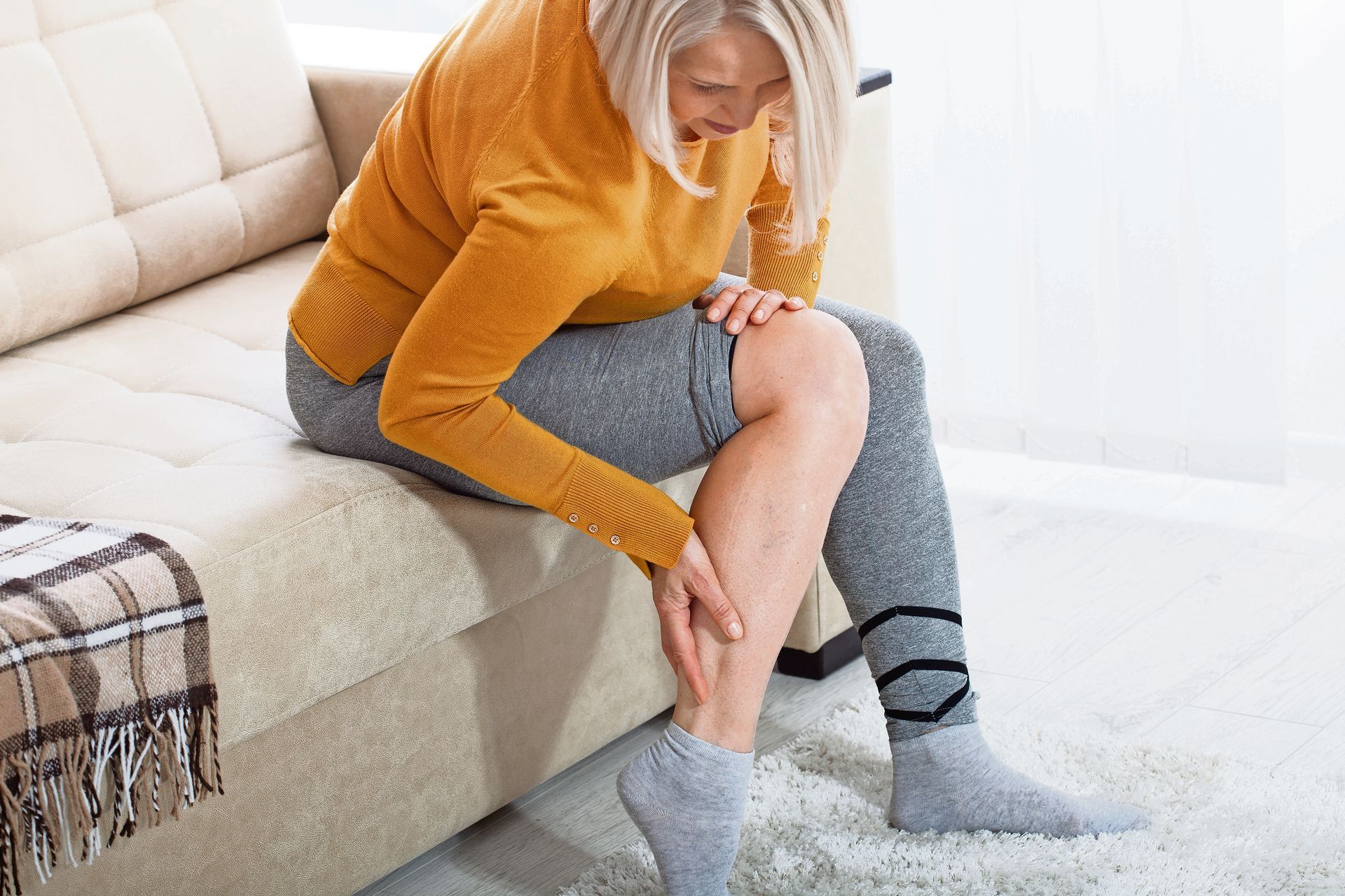 A person sitting on a sofa in a yellow sweater, holding their leg in apparent discomfort or pain.