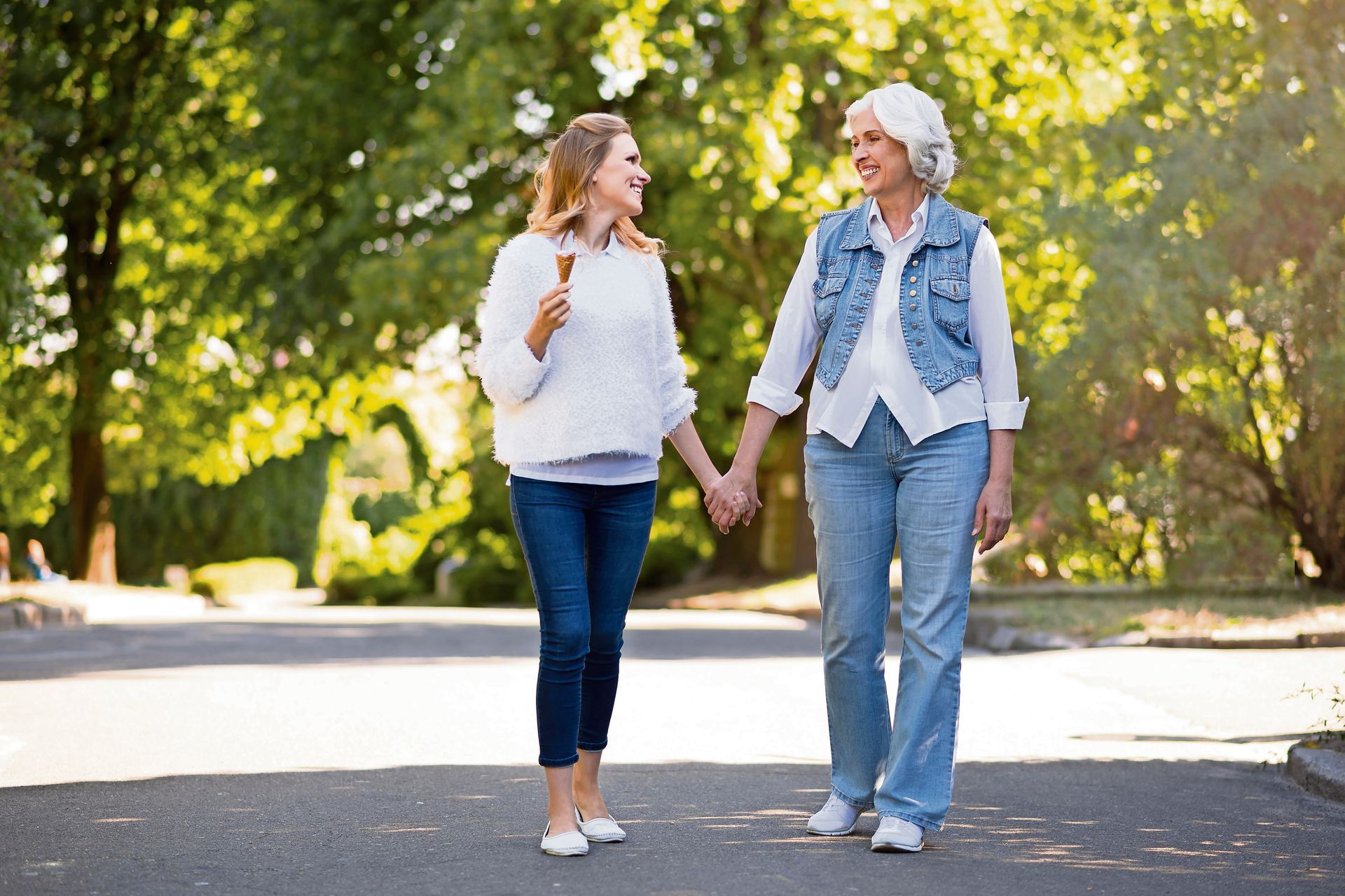 Two people walk hand-in-hand along a tree-lined path, smiling and talking while one holds a small wooden cross.