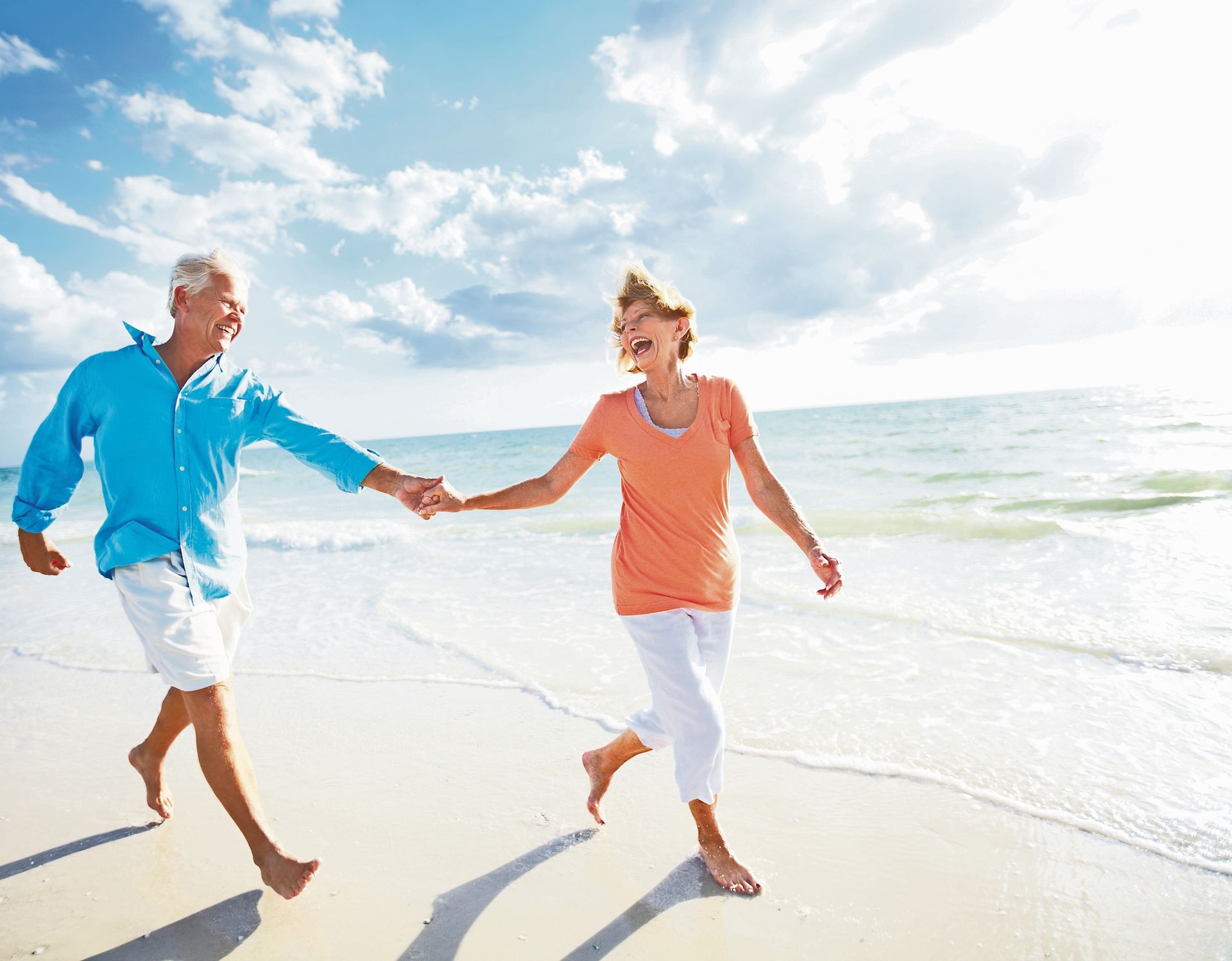 Two people holding hands and laughing while walking along a sunny beach near the ocean.