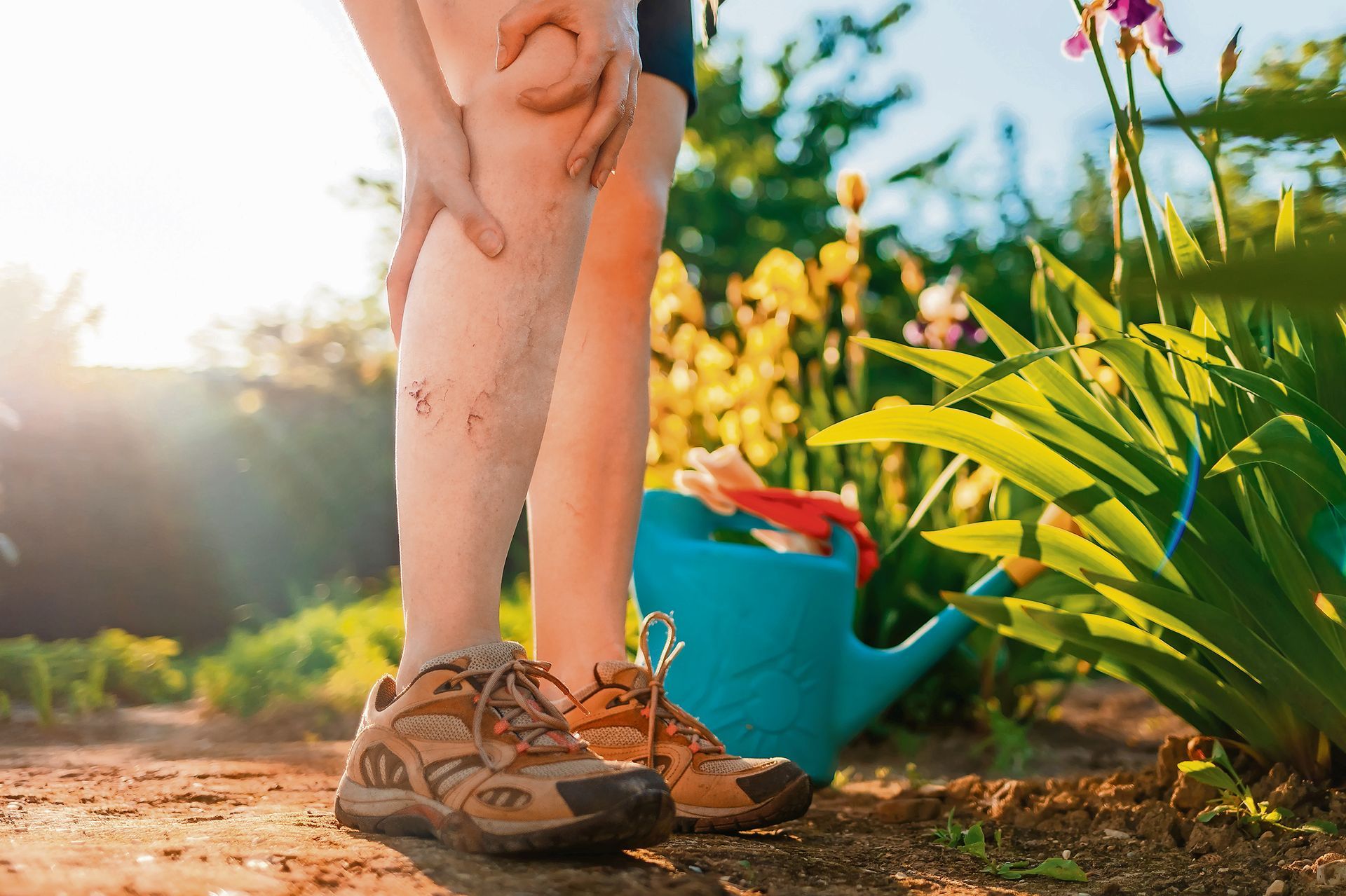 A person in a sunny garden stands holding their leg, which shows visible, dark-colored veins.