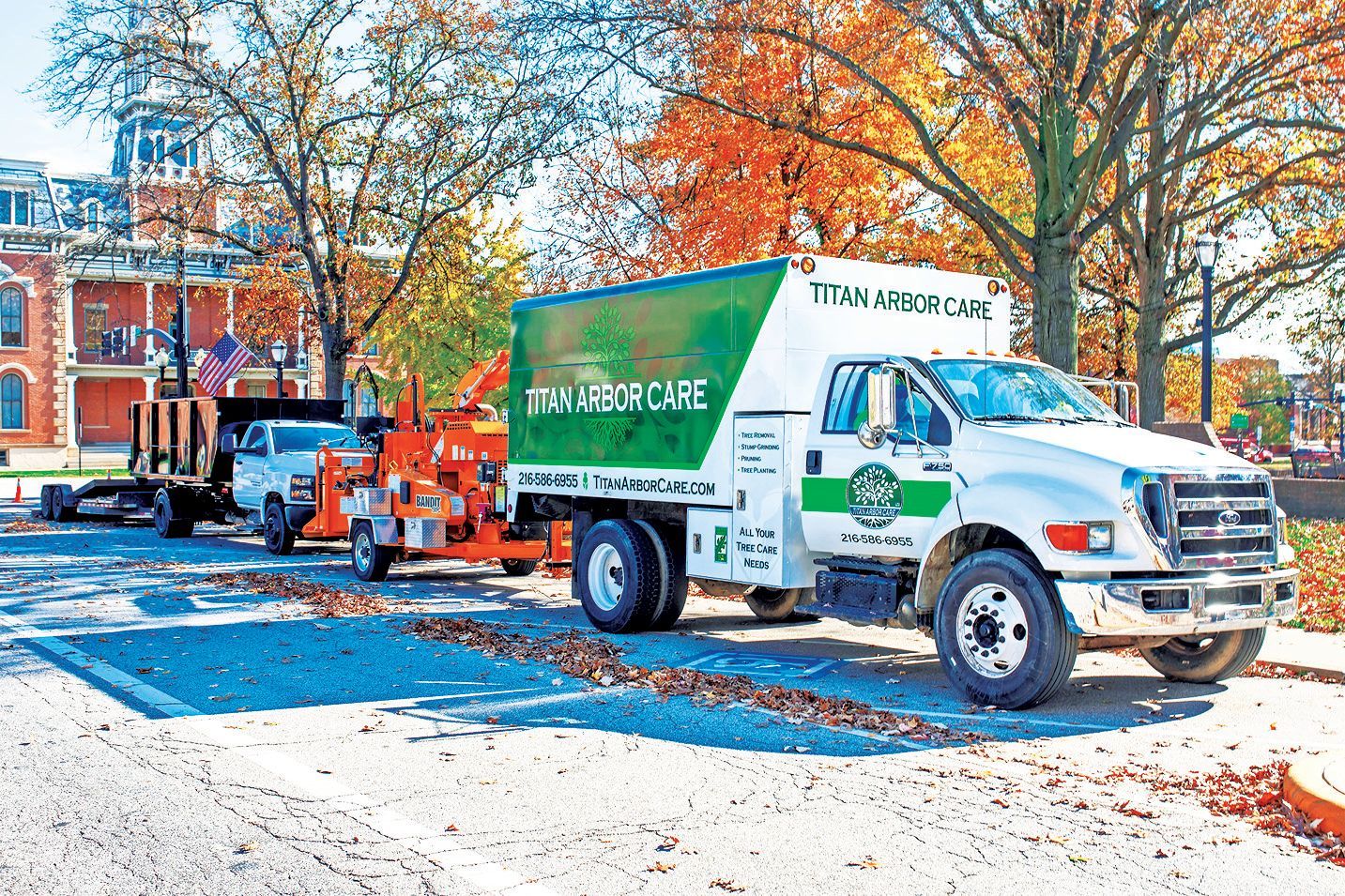 A white and green Arbor Care truck towing a wood chipper parked on a street lined with autumn trees and a brick building.