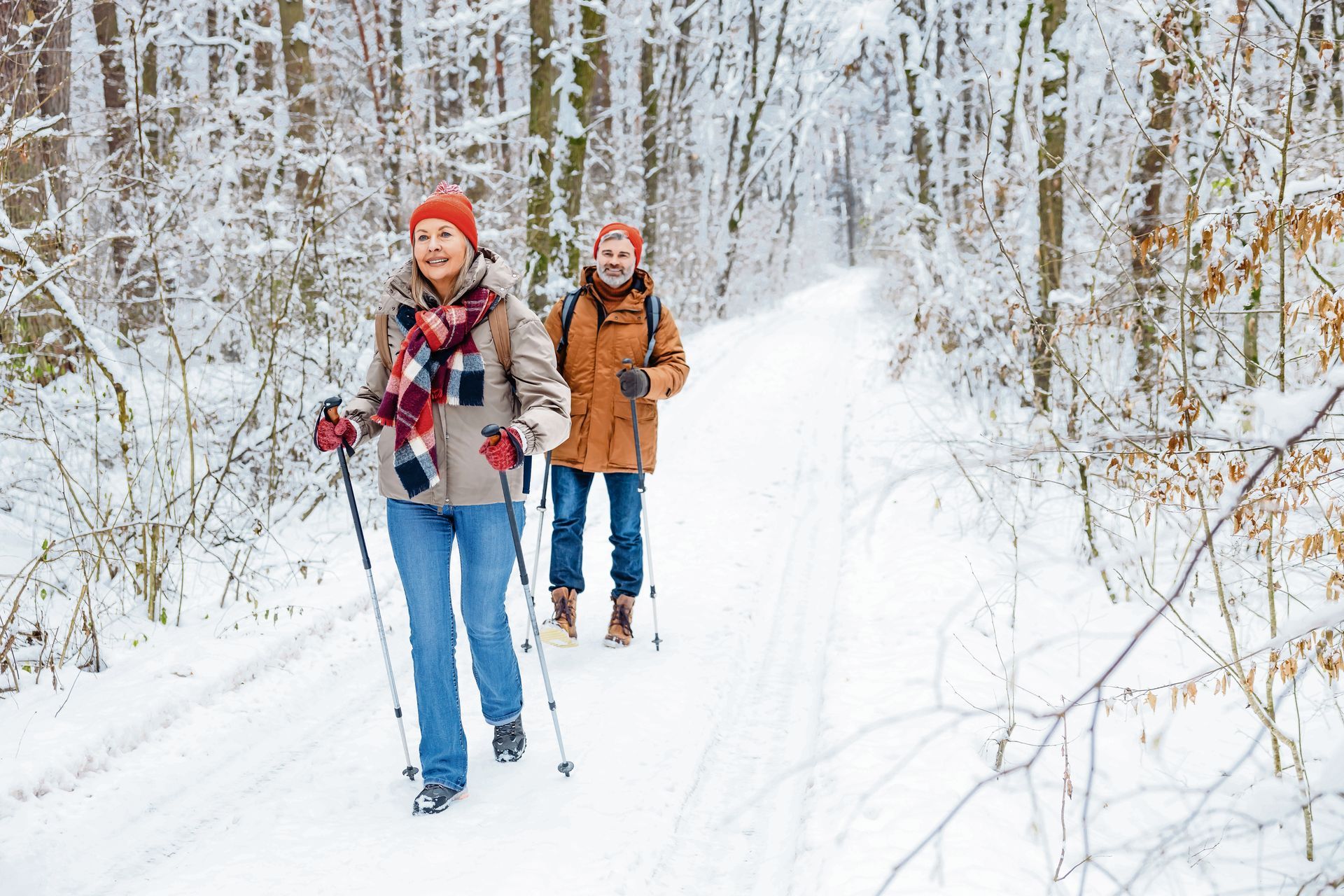 Two people in winter gear cross-country ski along a snow-covered path through a forest.