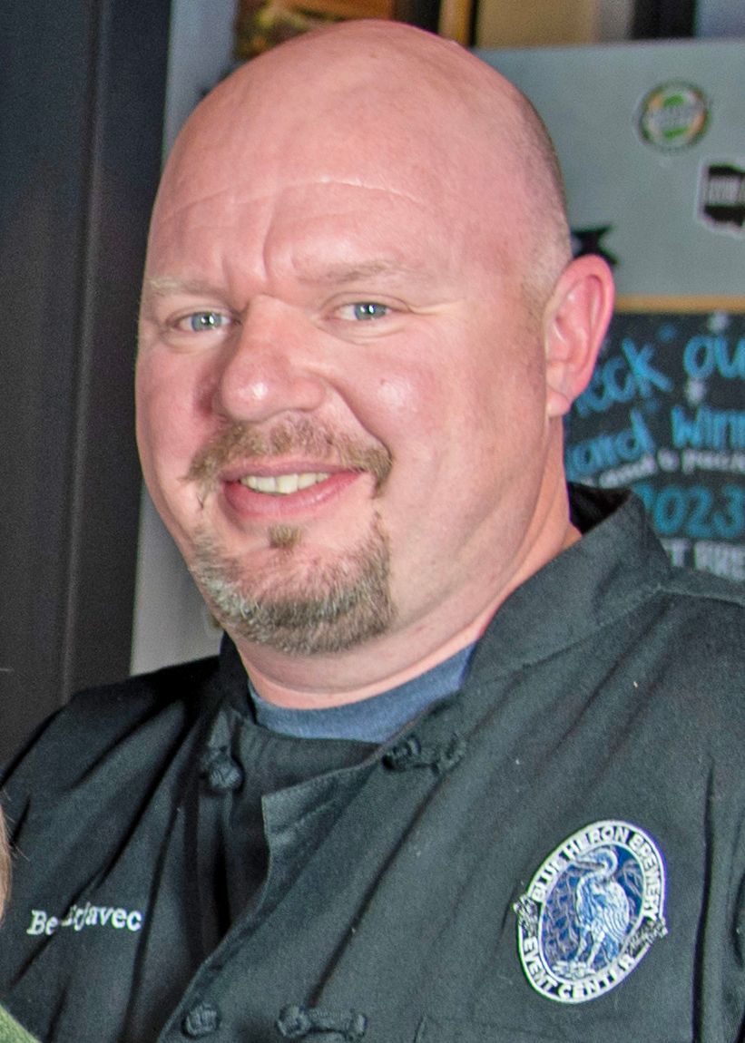 Smiling chef in a black uniform with a circular logo, standing in a kitchen setting.