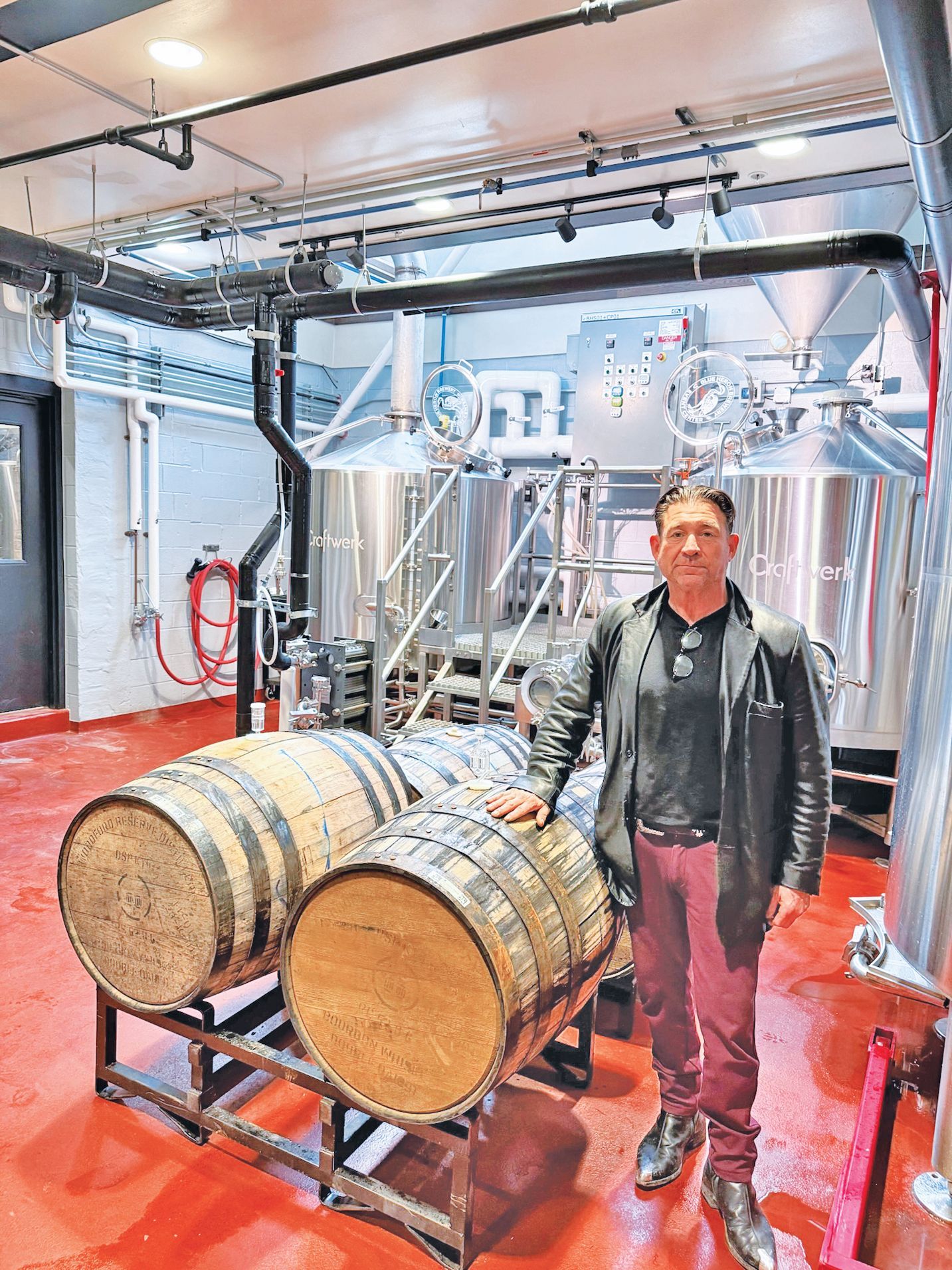 A man in a black jacket and reddish pants stands in a brewery next to two wooden barrels and stainless steel tanks.