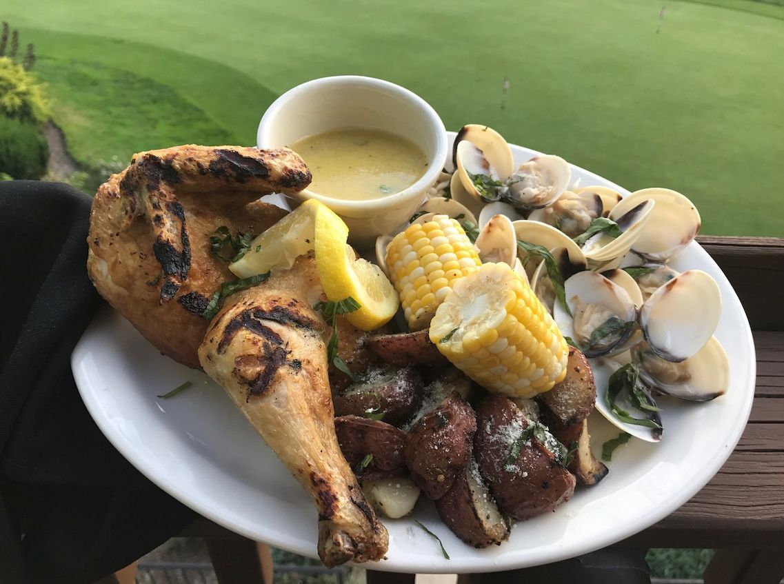 A plate of grilled chicken, clams, roasted potatoes, and corn on the cob with a dipping sauce, outdoors on a sunny day.