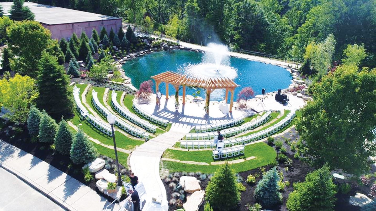 An aerial view of an outdoor wedding venue with circular seating facing a wooden pergola, a fountain, and a pond.