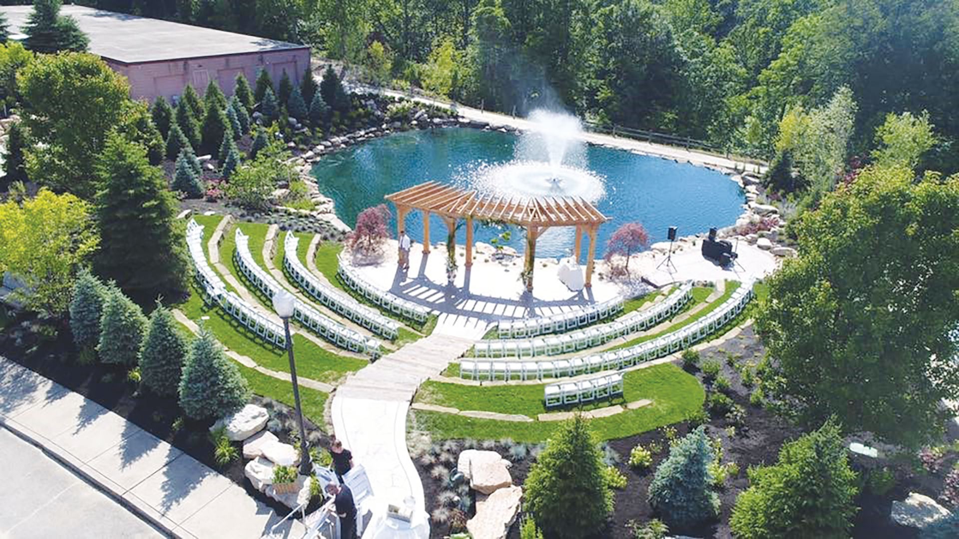 An outdoor wedding venue with rows of white chairs facing a wooden pergola, a circular pond, and a large fountain.