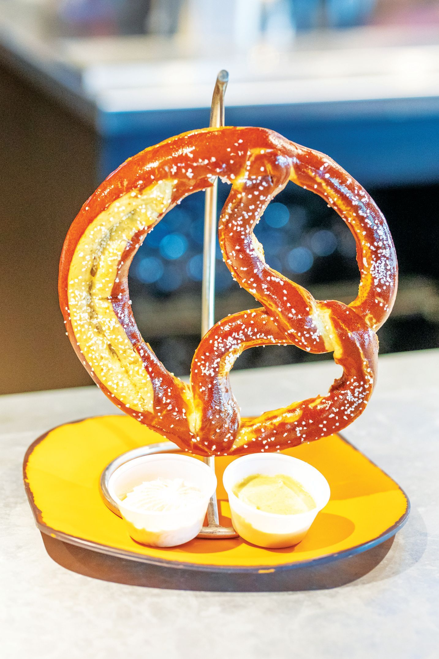 A large, salted pretzel hangs on a metal stand above two small cups of dipping sauce on a yellow tray.