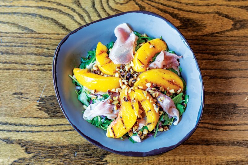 A grilled peach and prosciutto salad topped with chopped nuts, served in a blue bowl on a dark wooden table.