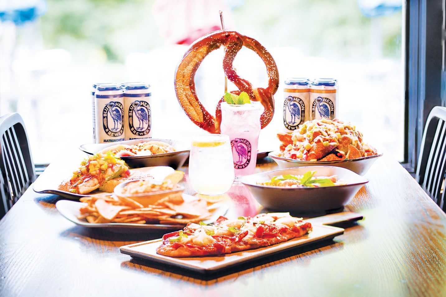 A table full of pub food and drinks, including a large pretzel, flatbread, chips, and canned beer, in a bright setting.