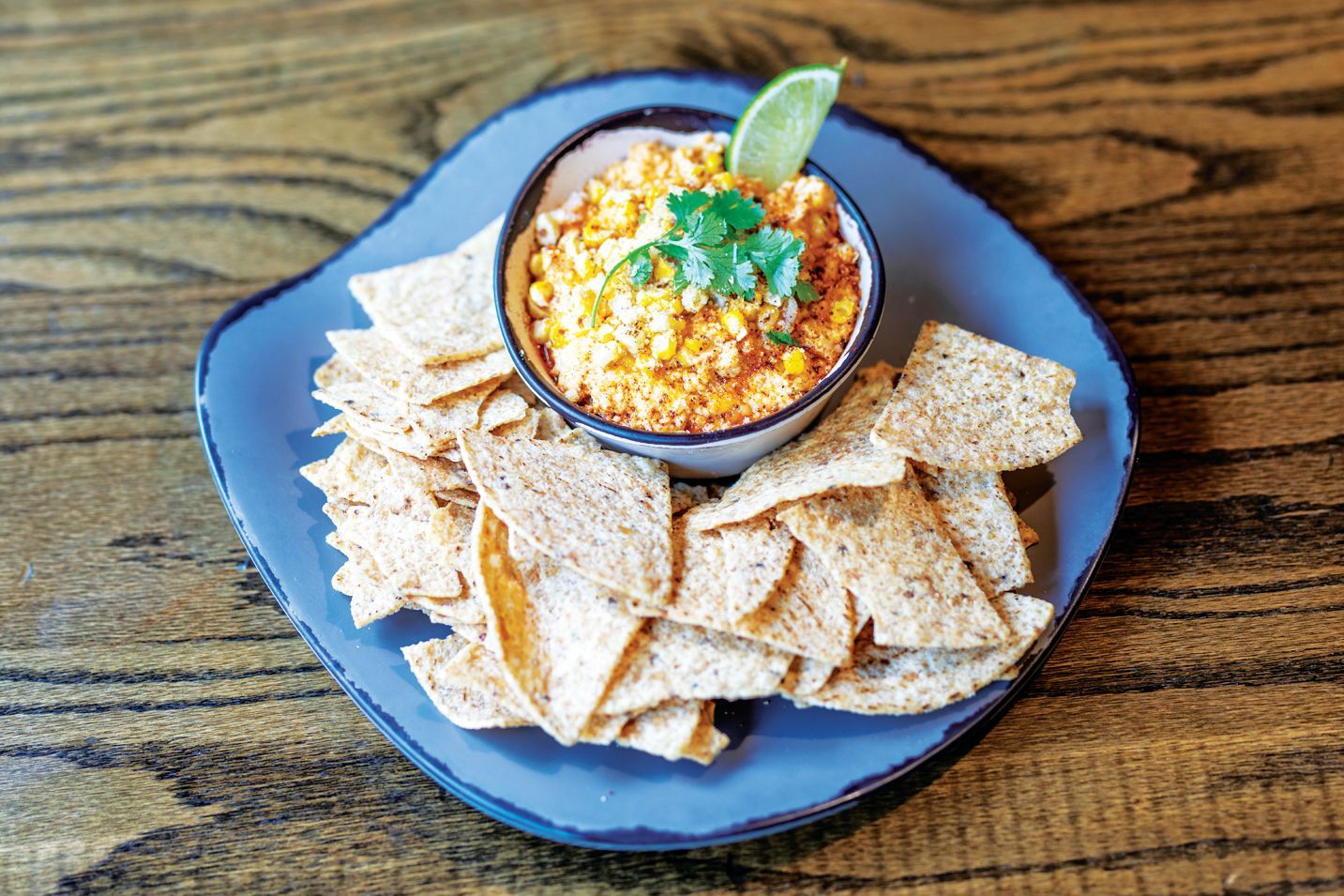 A bowl of elote dip topped with cilantro and a lime wedge, served with tortilla chips on a blue plate on a wooden table.