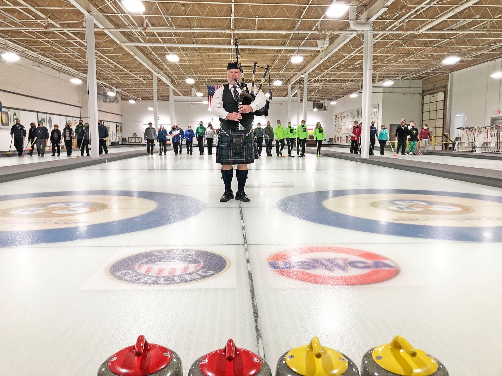 A person in traditional Scottish attire plays bagpipes on a curling rink, with curling stones in the foreground.