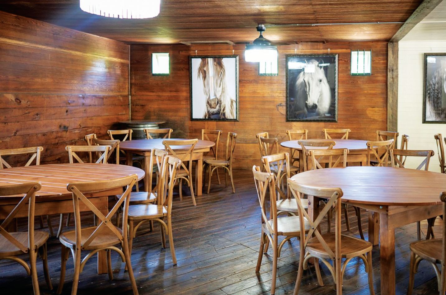 Rustic dining room with wood-paneled walls, round wooden tables, cross-back chairs, and framed horse artwork.