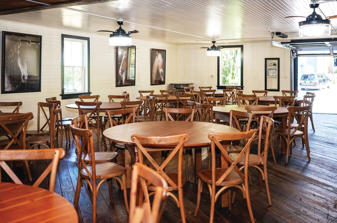 An indoor dining room with round wooden tables, cross-back chairs, ceiling fans, and framed art on white walls.