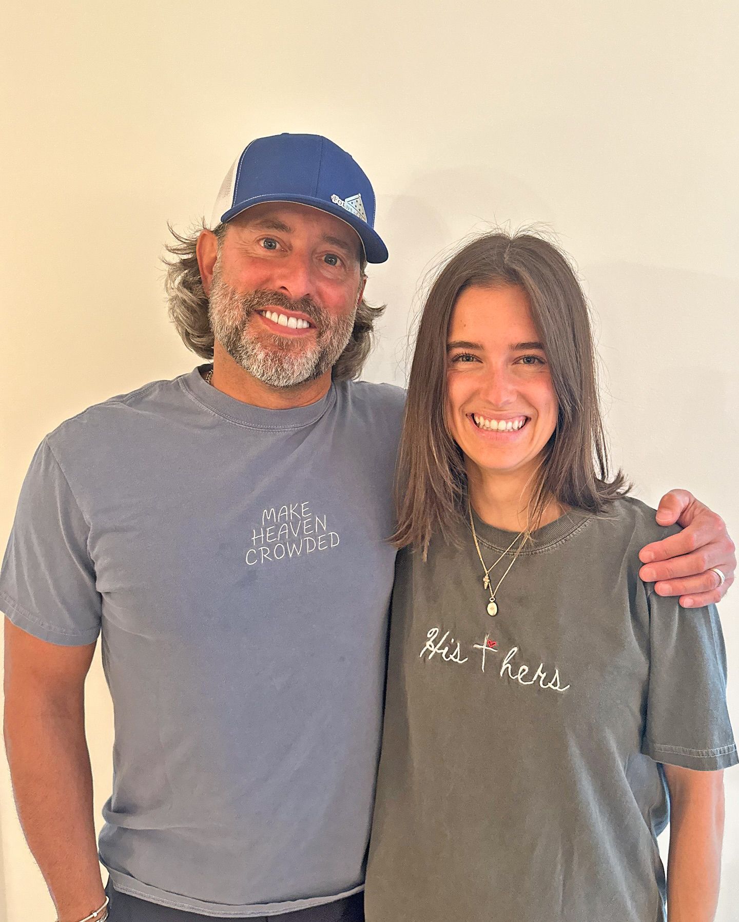 A man in a blue cap and t-shirt smiling with an arm around a woman wearing a dark grey graphic t-shirt and necklace.