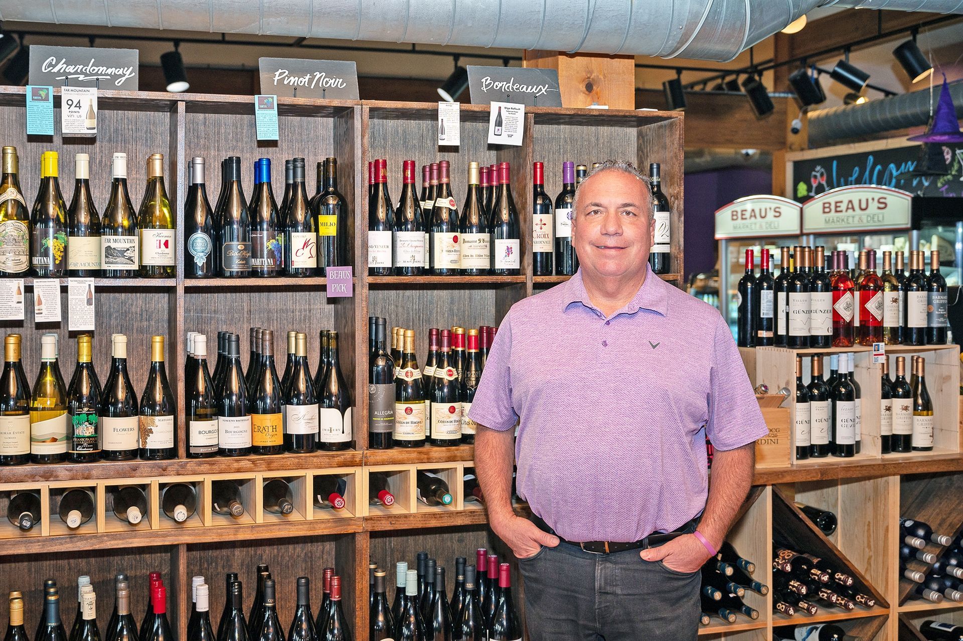 A smiling individual in a purple button-down shirt standing in a wine shop with shelves of bottles in the background.